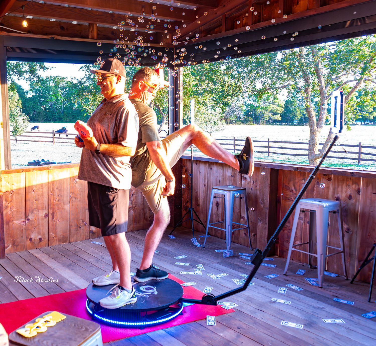 Two people stand on a balance board trainer at an outdoor covered deck with bar stools and string lights.