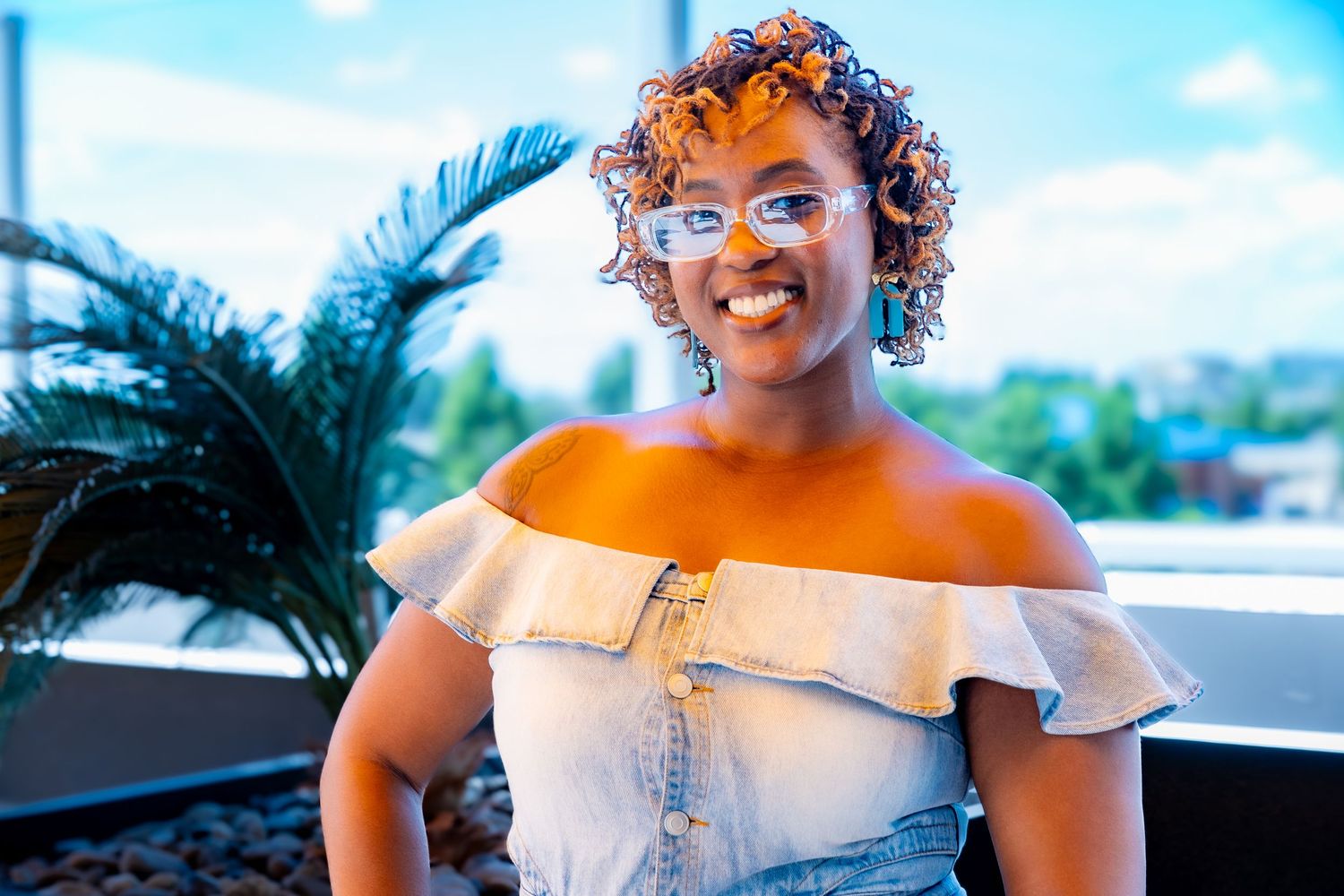 Person in light denim off-shoulder dress posing outdoors against blue sky.