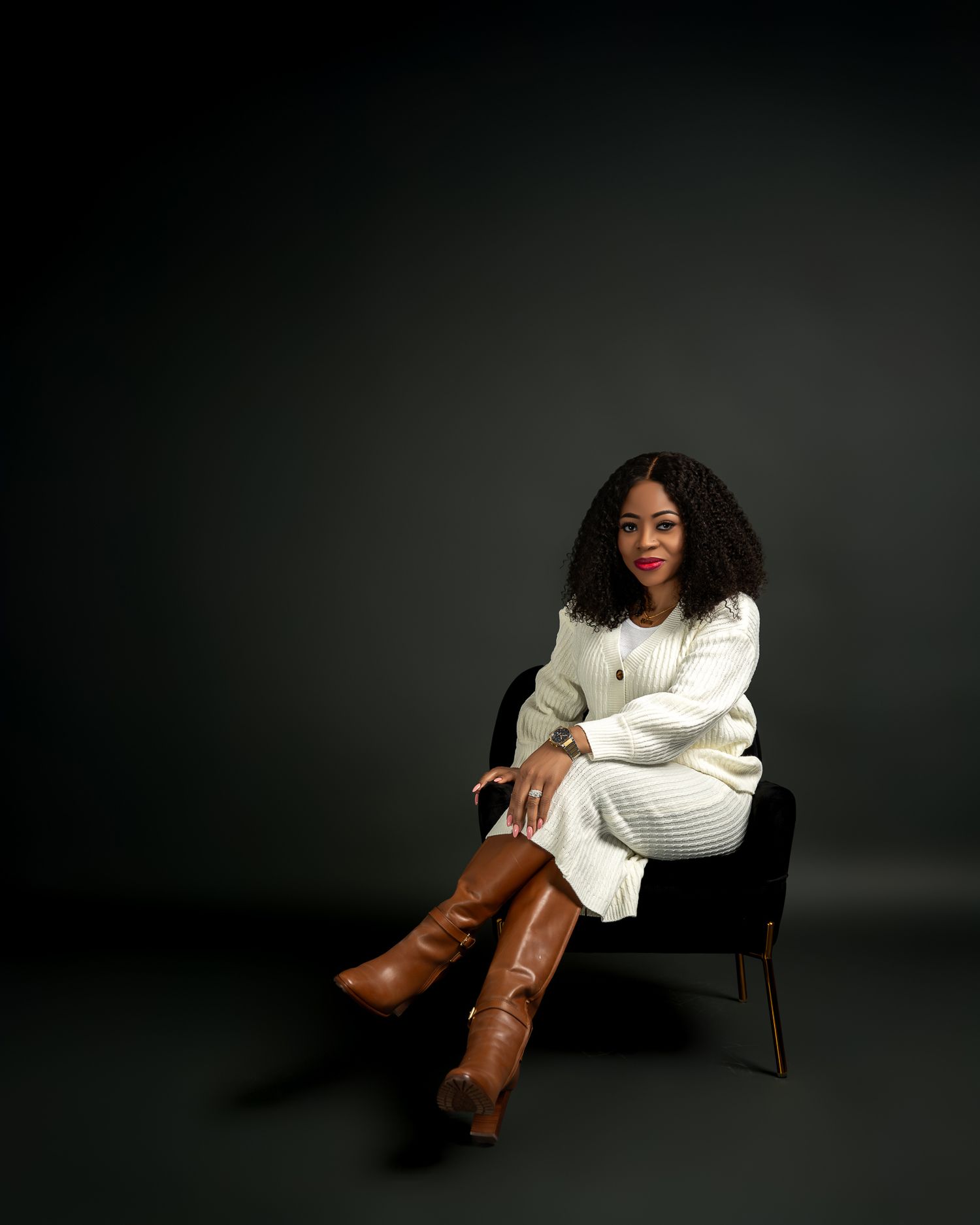 Person in white sweater and brown boots poses elegantly on chair against dark backdrop.