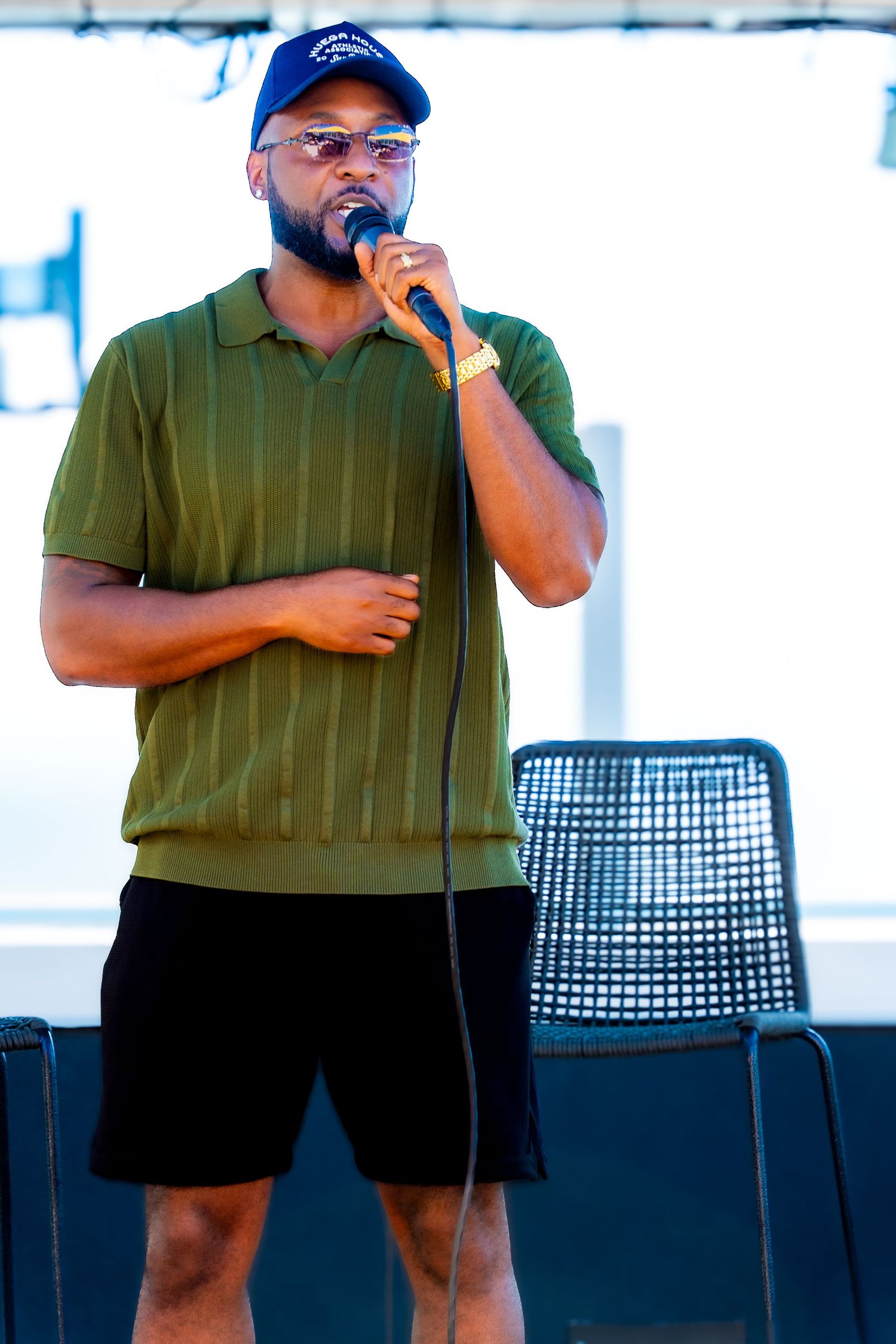 Performer in a green polo and baseball cap singing into a microphone on stage.