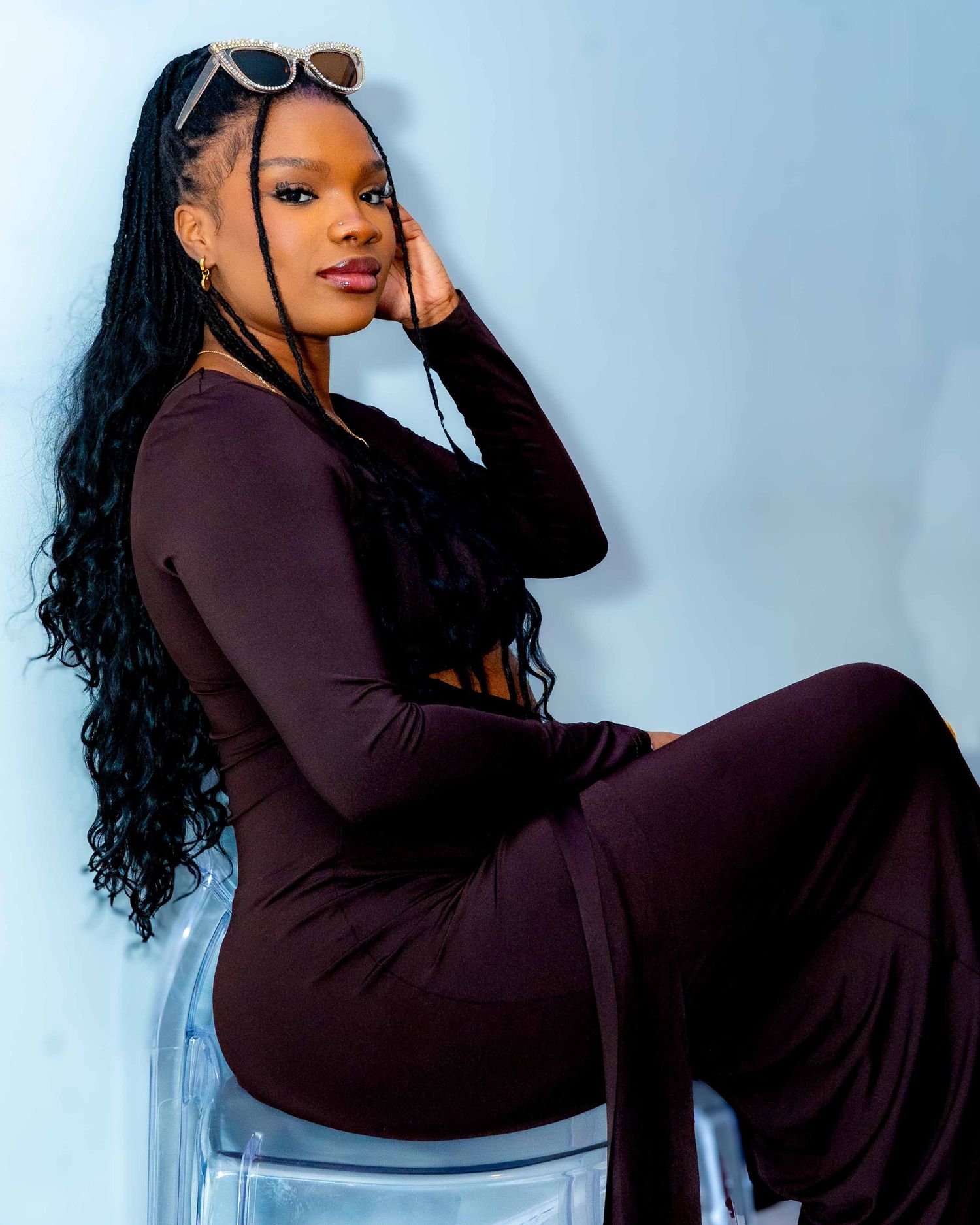 Model wearing burgundy outfit poses artistically on clear chair during studio photoshoot.