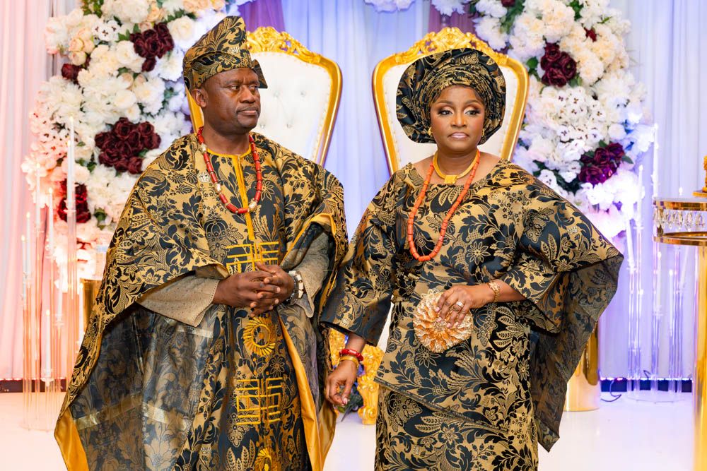 Couple in matching black and gold traditional formal wear pose by floral wedding backdrop.
