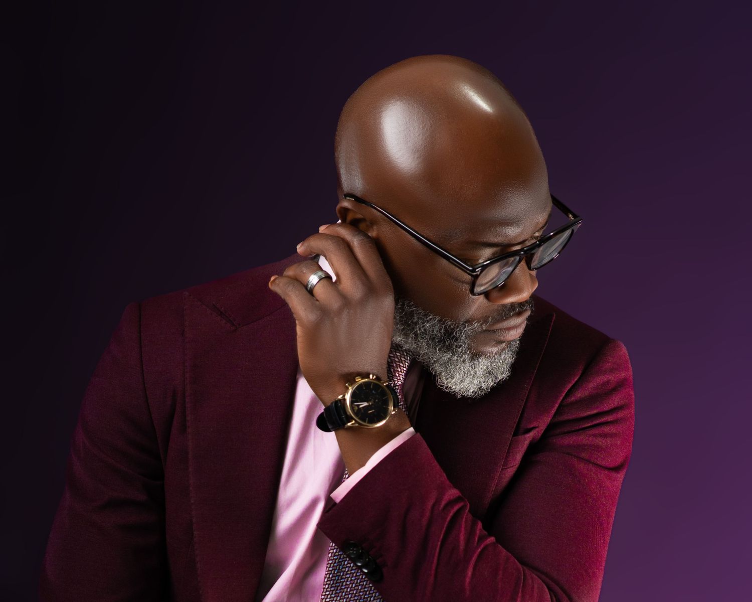 Professional studio portrait of person in burgundy blazer and pink shirt wearing glasses and gold watch against dark background.