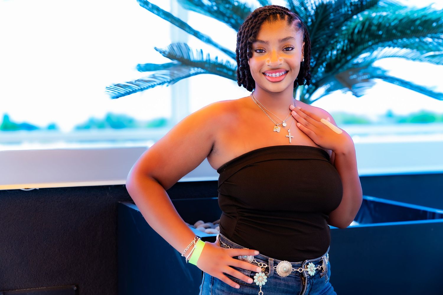 Stylish woman in black top and jeans poses with palm trees in background.