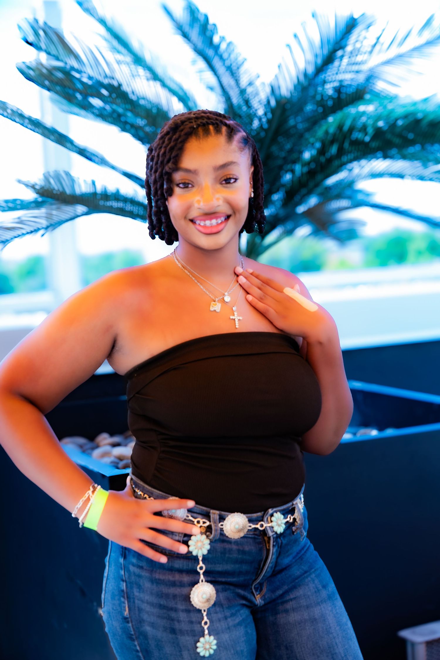 Woman in black strapless top and jeans poses with decorative belt against palm trees.