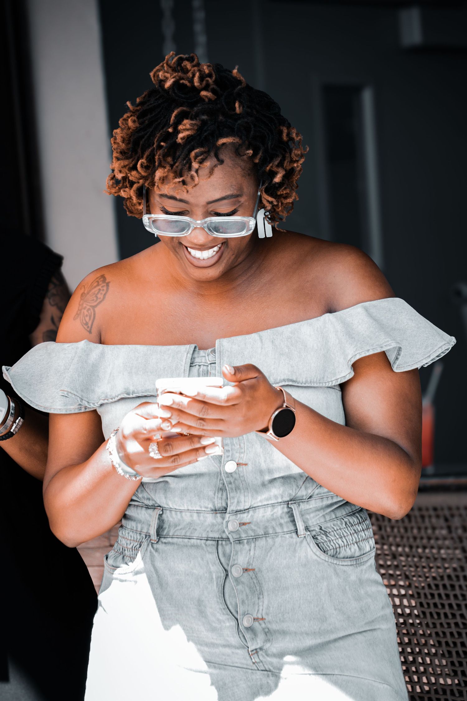 Person wearing light denim off-shoulder dress and glasses smiling while posing against dark background.