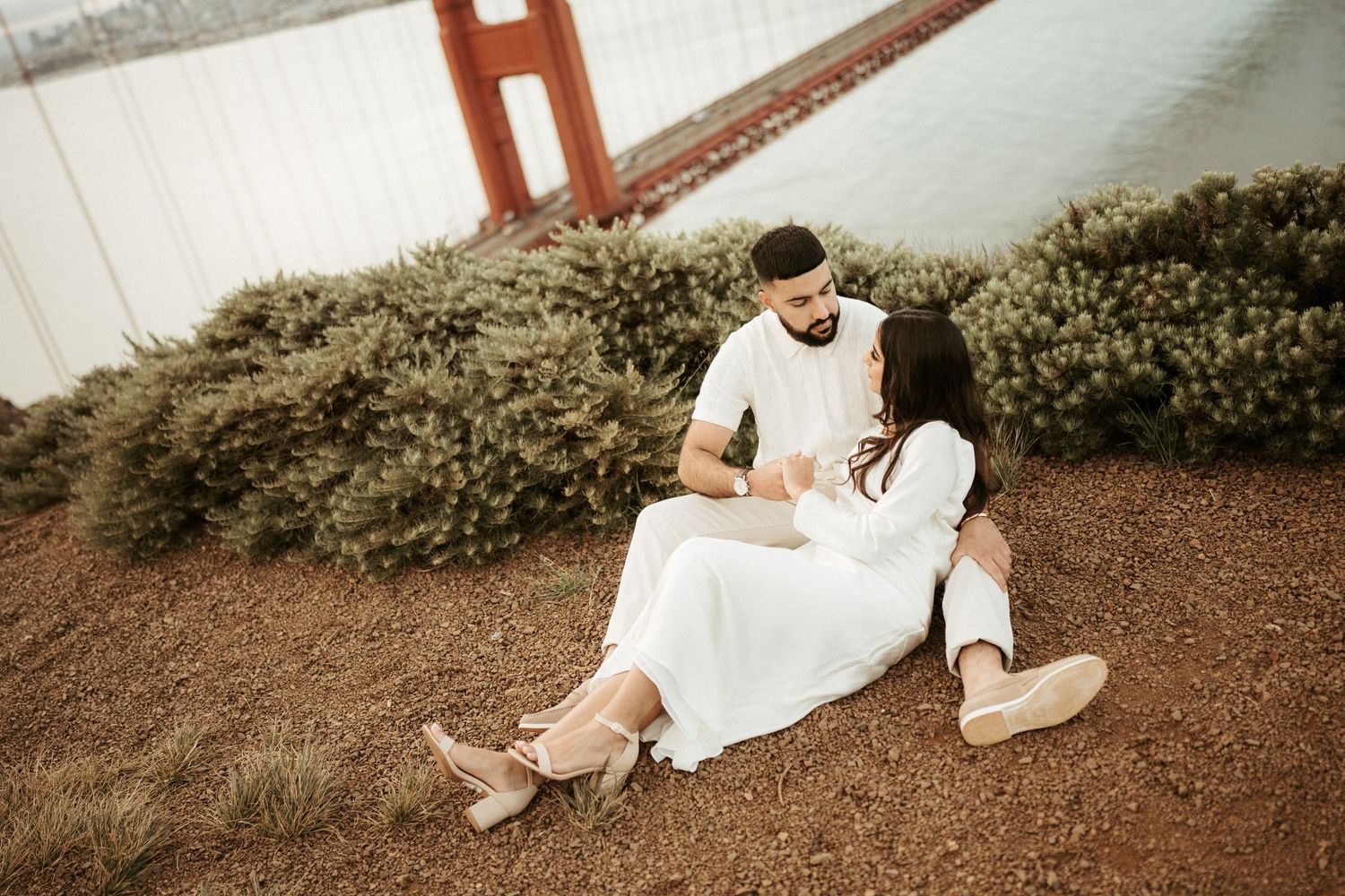 A couple in white attire poses together on a rustic wooden bridge during their wedding photoshoot.