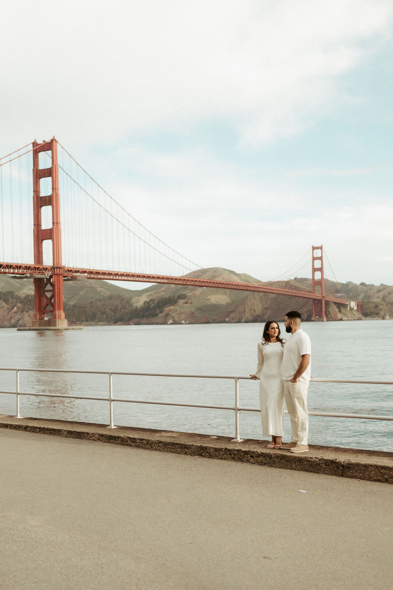 A couple in white outfits stands along the waterfront with the iconic Golden Gate Bridge in San Francisco behind them.