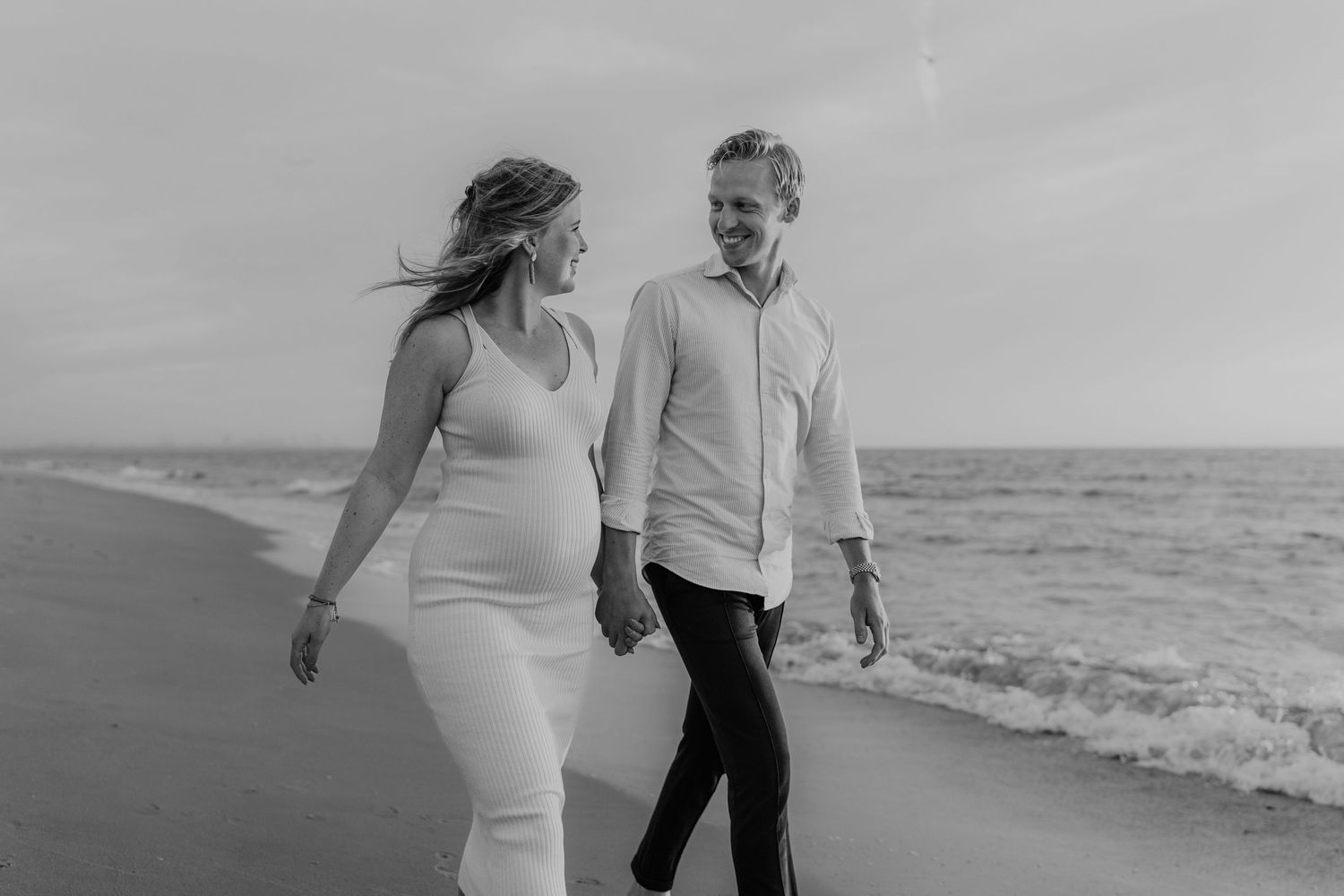 Een zwanger koppel op het strand dat gelukkig naar elkaar kijkt tijdens een zwangerschapsshoot tijdens het gouden uur aan zee. Zwart-wit.