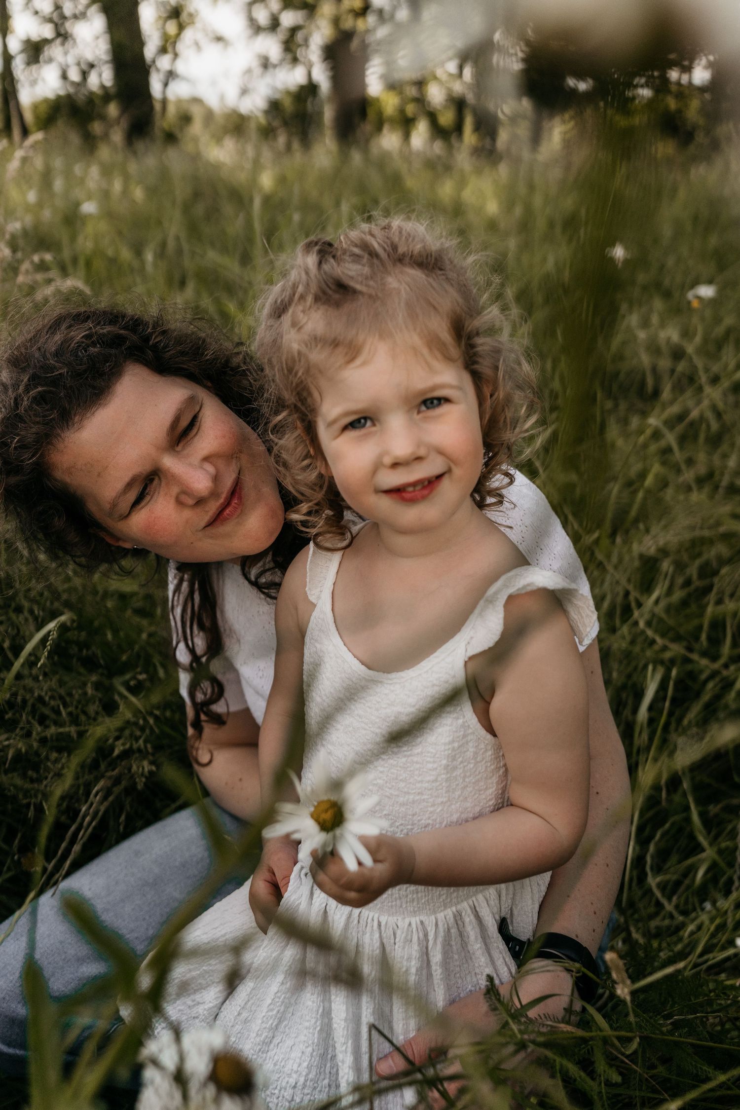 Moeder knuffelt lachend haar dochter terwijl dochter in de camera kijkt tijdens een fotoshoot in een bloemenveld in Goirle