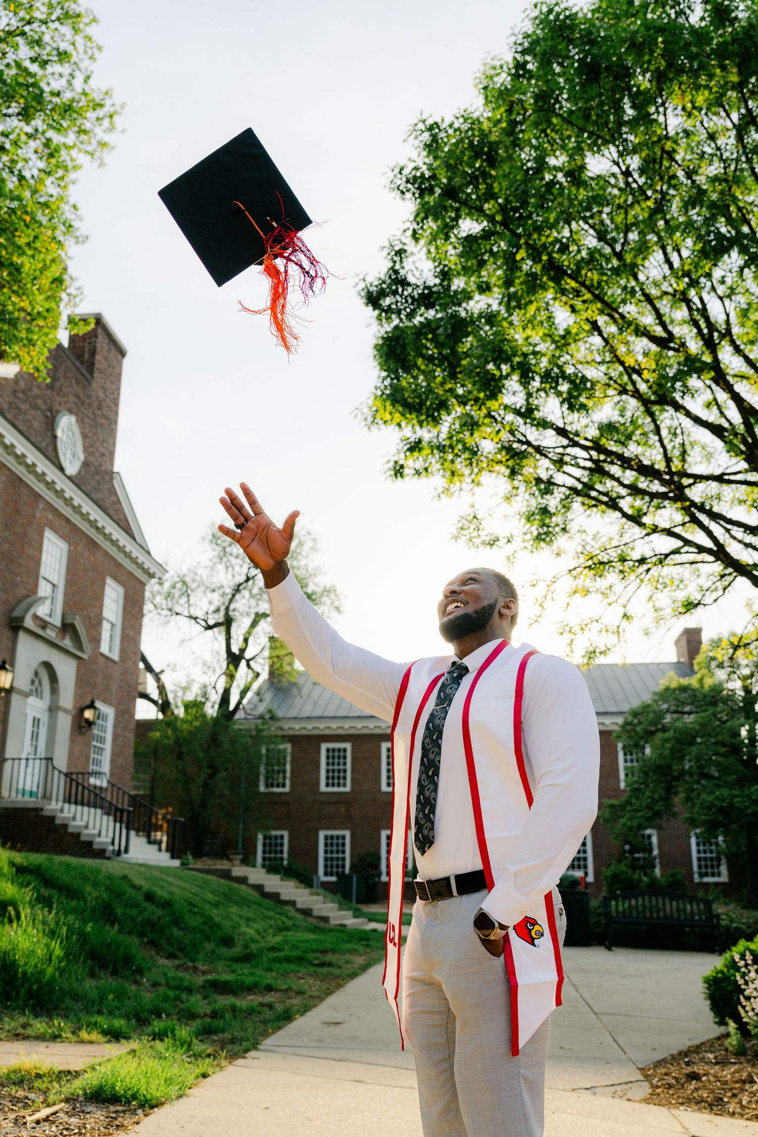 College graduate in white cap and gown celebrating on campus with brick buildings in background.