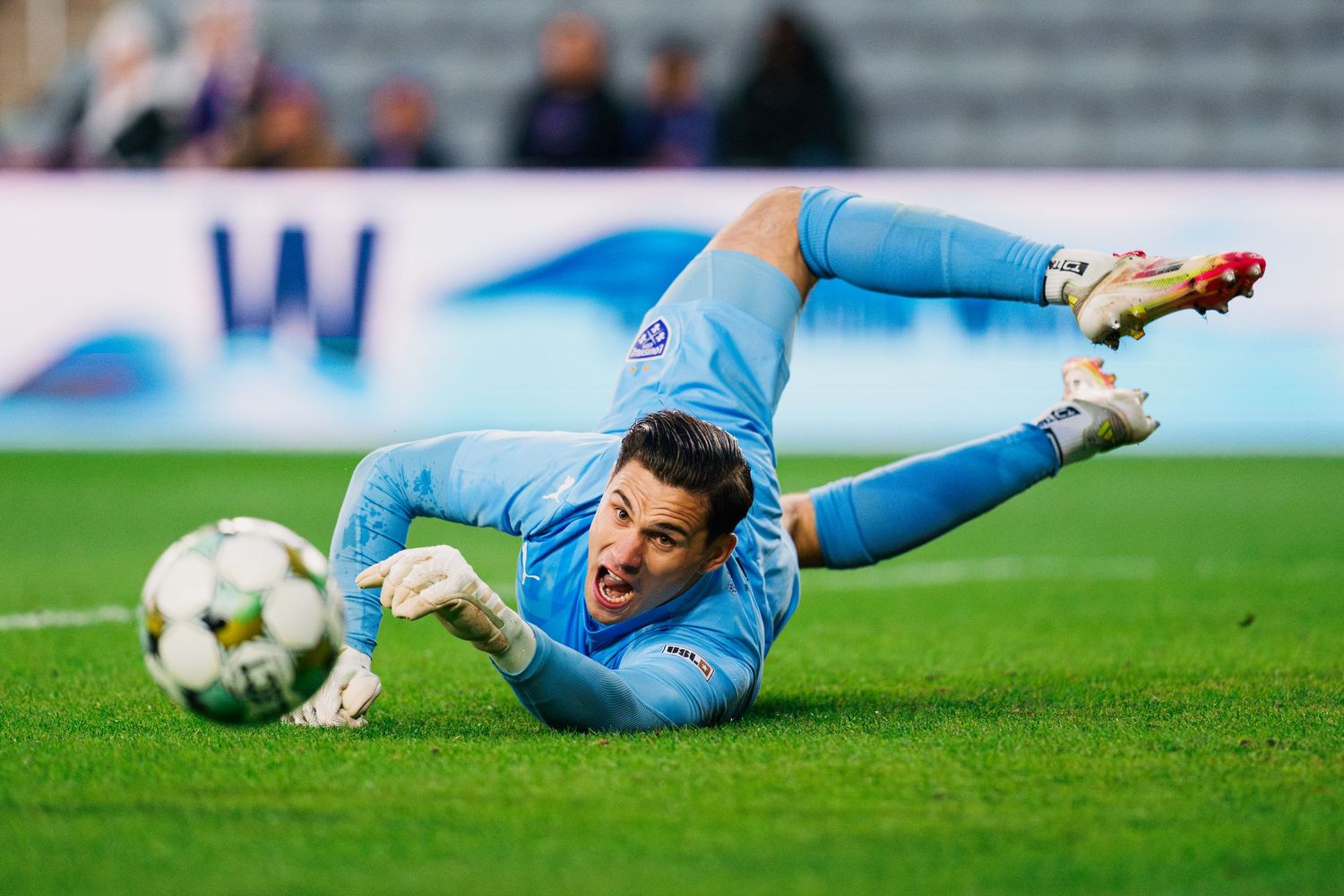 A goalkeeper in light blue uniform dives to make a save during a soccer match on a grass field.