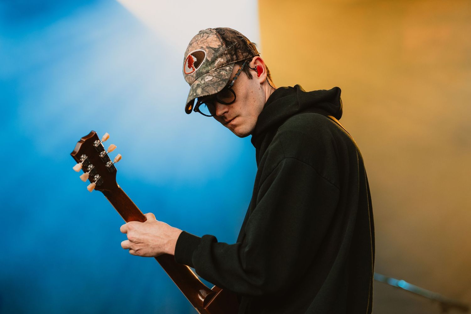 Person in black hoodie playing guitar against blue and orange backdrop.
