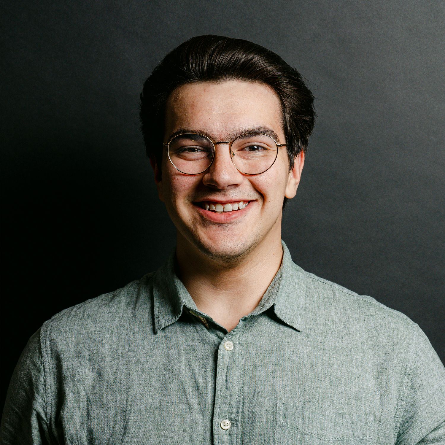 Professional headshot against dark background wearing grey button-up shirt.