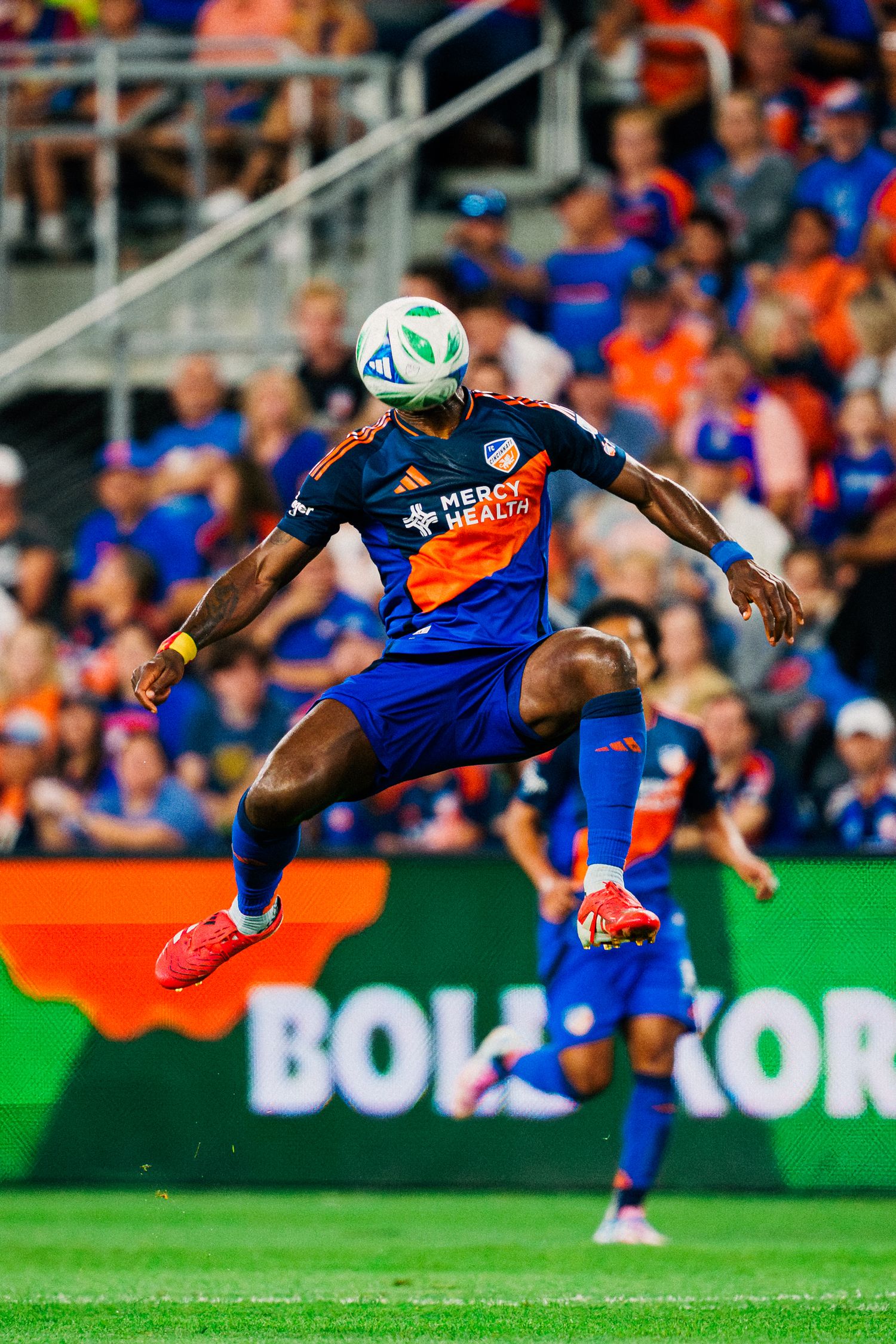 Soccer player in blue uniform performs acrobatic bicycle kick during match on green field with crowd in background.