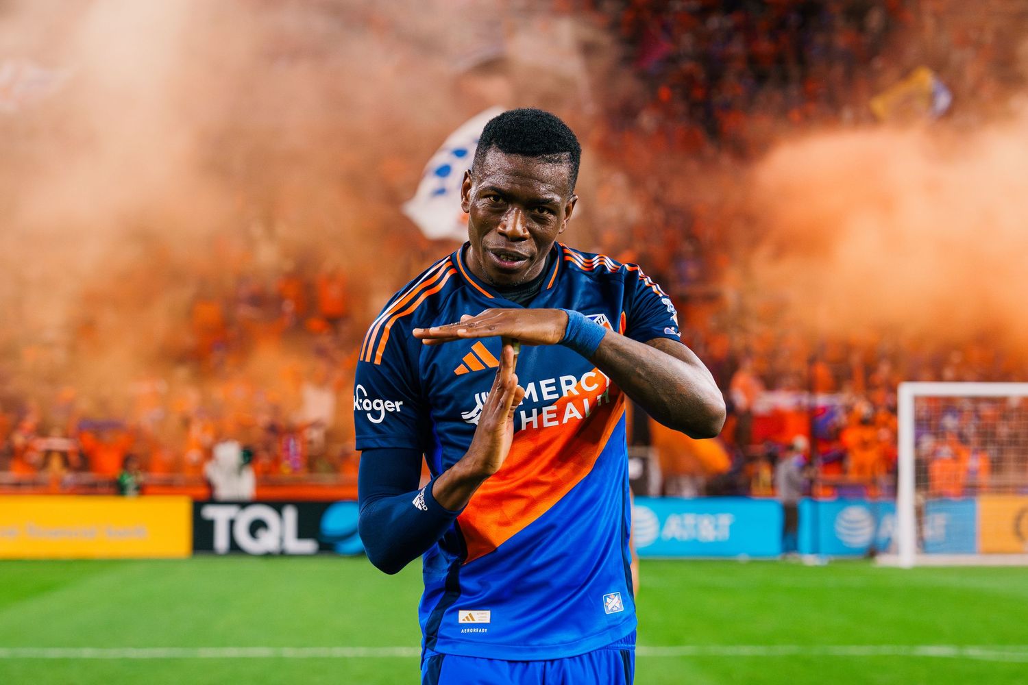 Soccer player celebrates scoring a goal with teammates during a match as orange smoke fills the stadium.
