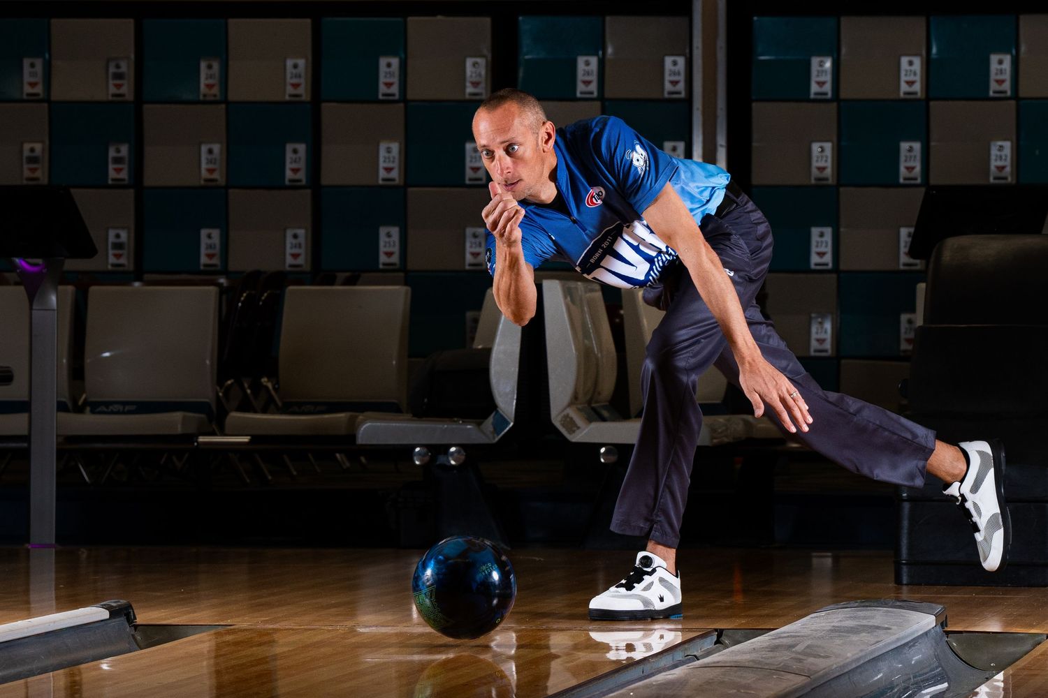 A bowler in blue attire performs a sliding delivery on a polished bowling alley lane.