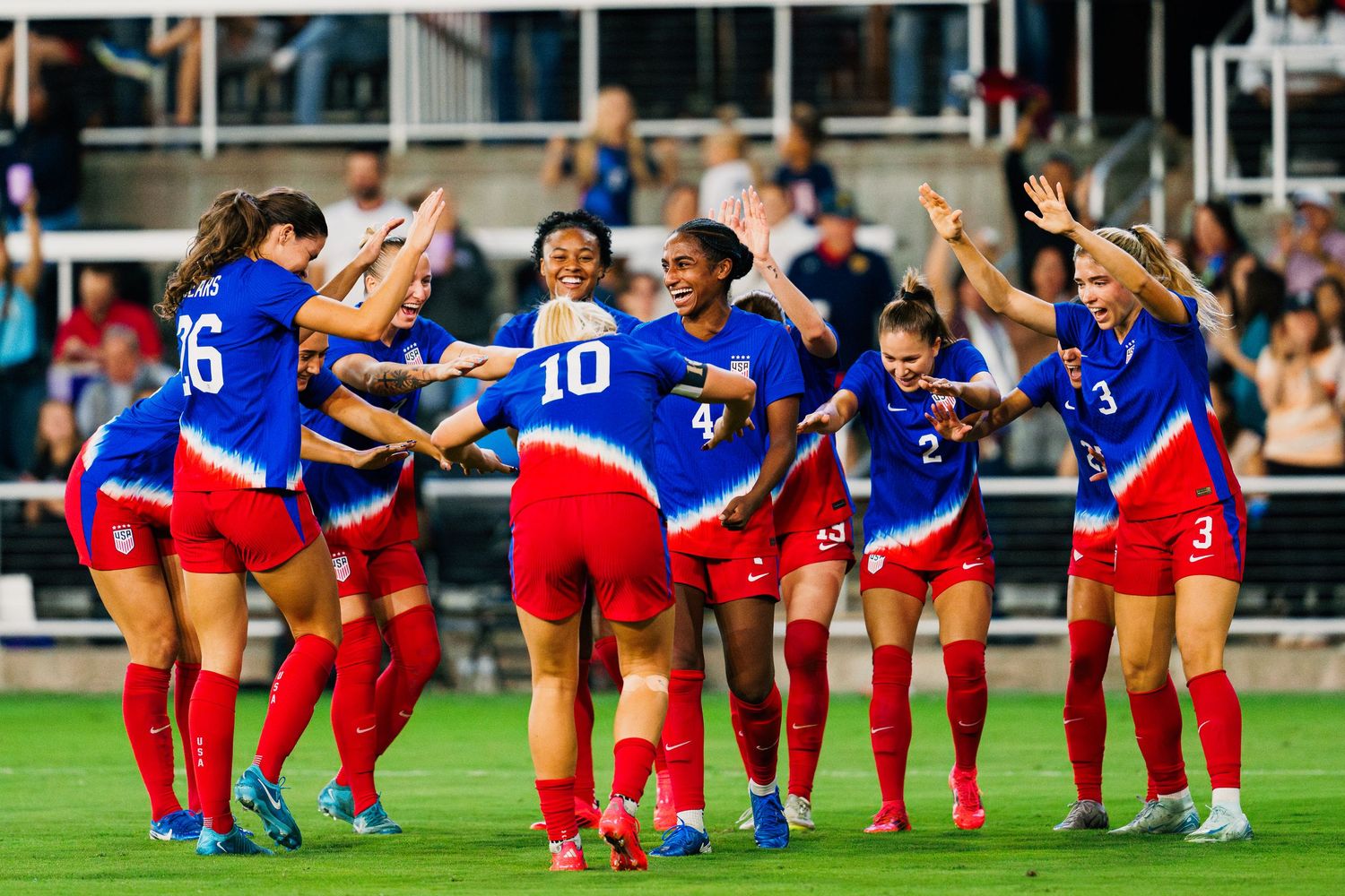 Soccer players in red and blue uniforms perform a synchronized celebration wave on the field during a match.