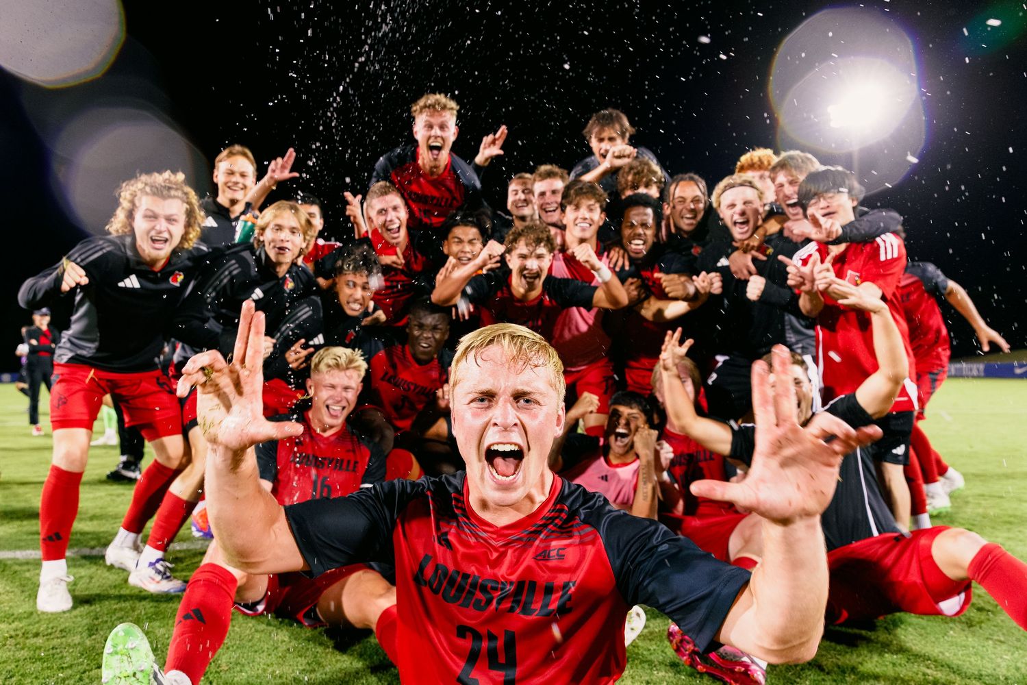Rugby players in red uniforms celebrate on the field at night after winning a match.