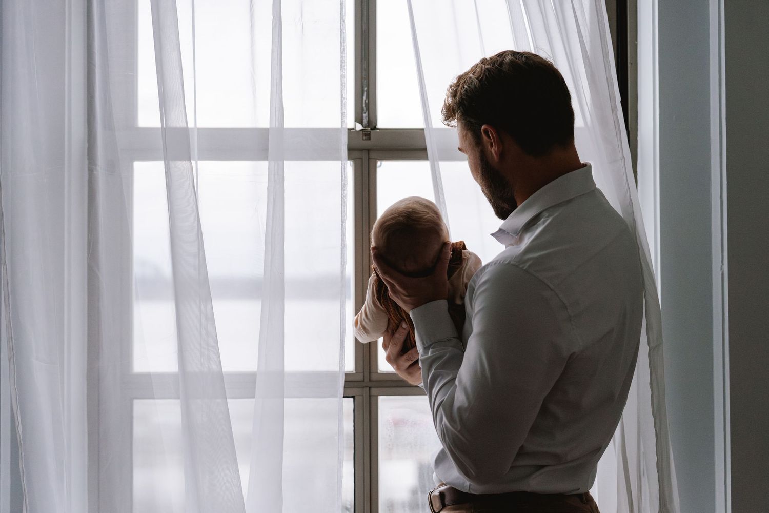 Two people embrace in silhouette against a bright window with white curtains in an emotional moment.