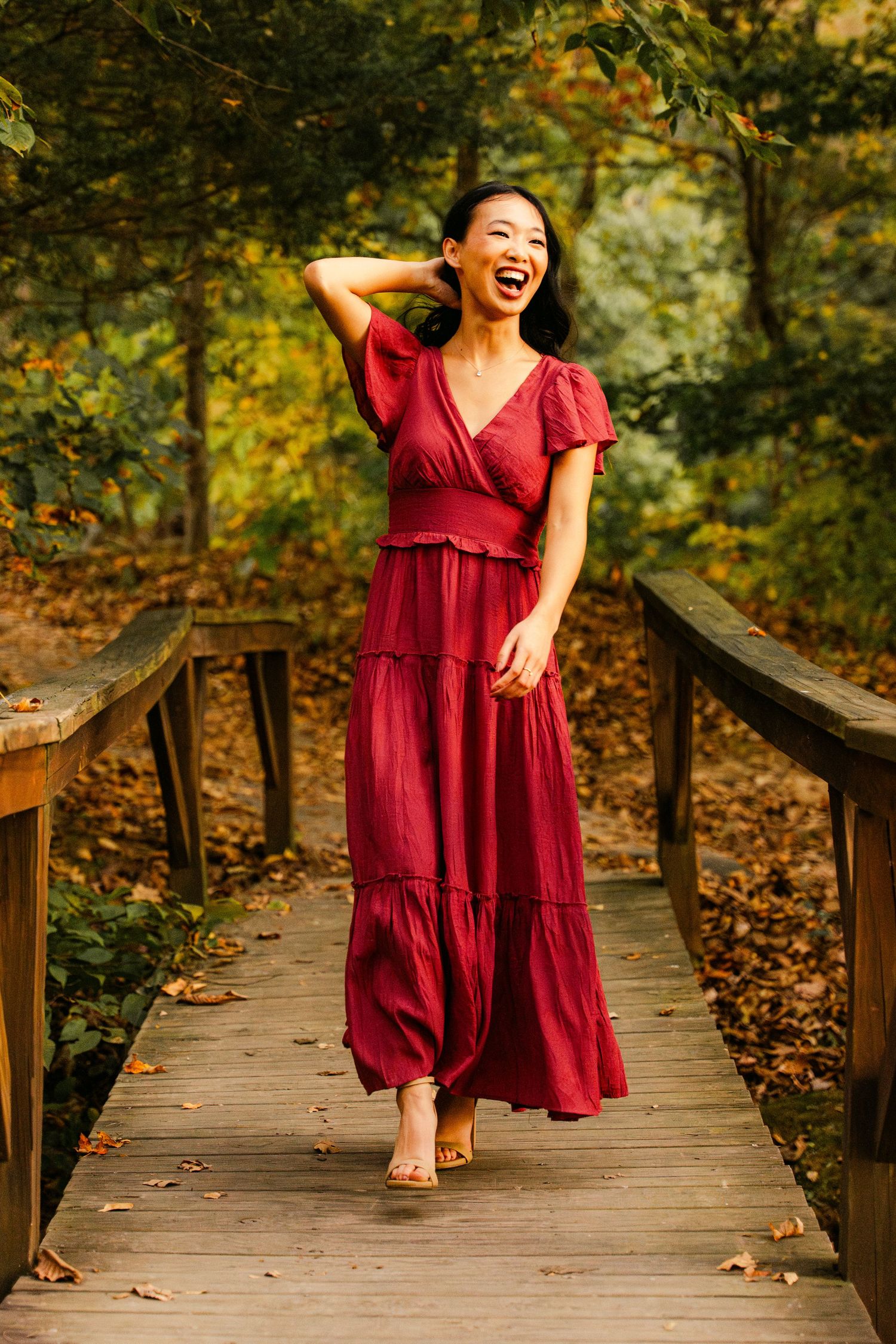 Person in burgundy dress twirling on wooden boardwalk surrounded by fall foliage.