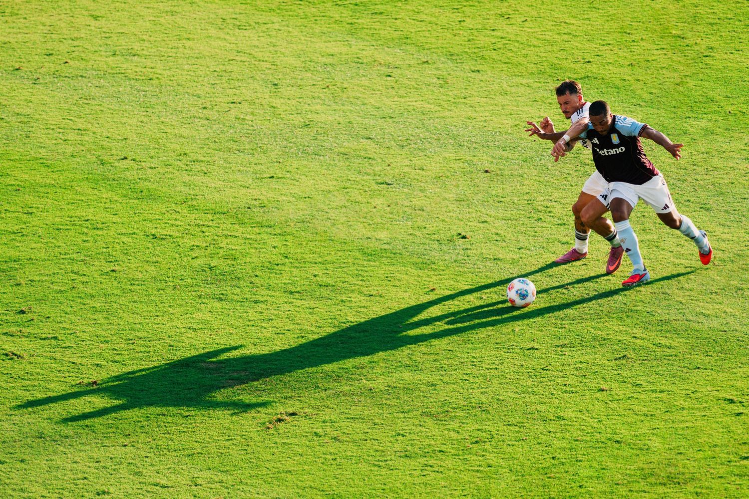 Soccer player dribbles ball across bright green grass field casting long shadow in afternoon sunlight.