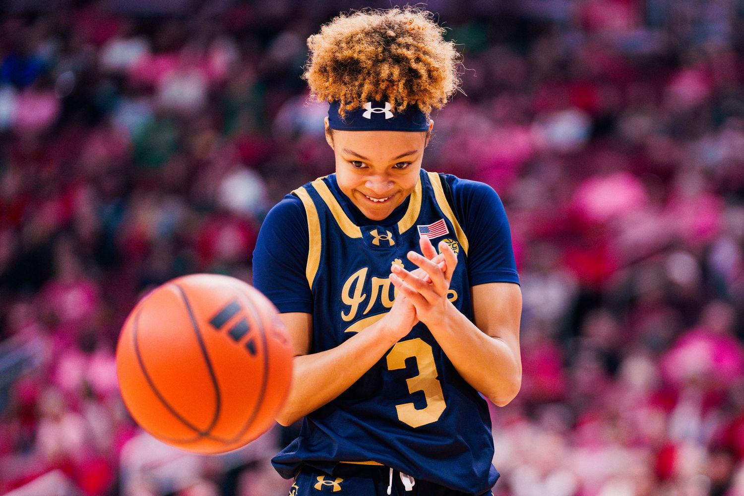 A basketball player in a navy blue jersey number 3 dribbles during a game with a blurred pink background.