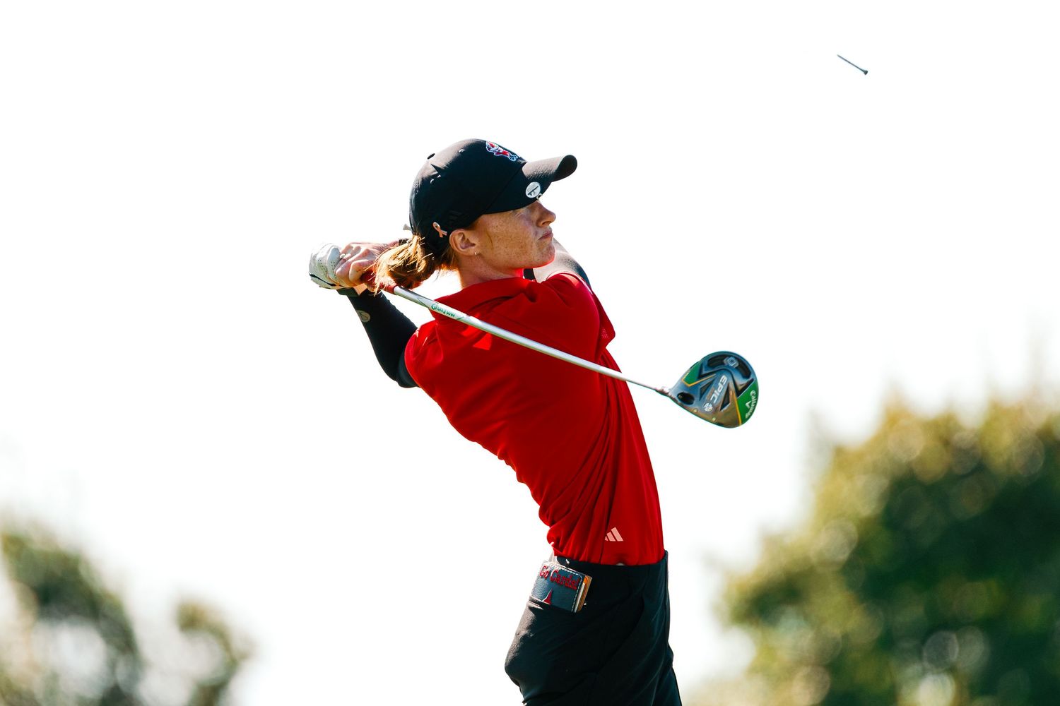 A golfer in a red sweater and black cap swings a driver during a golf shot against a blurred background.