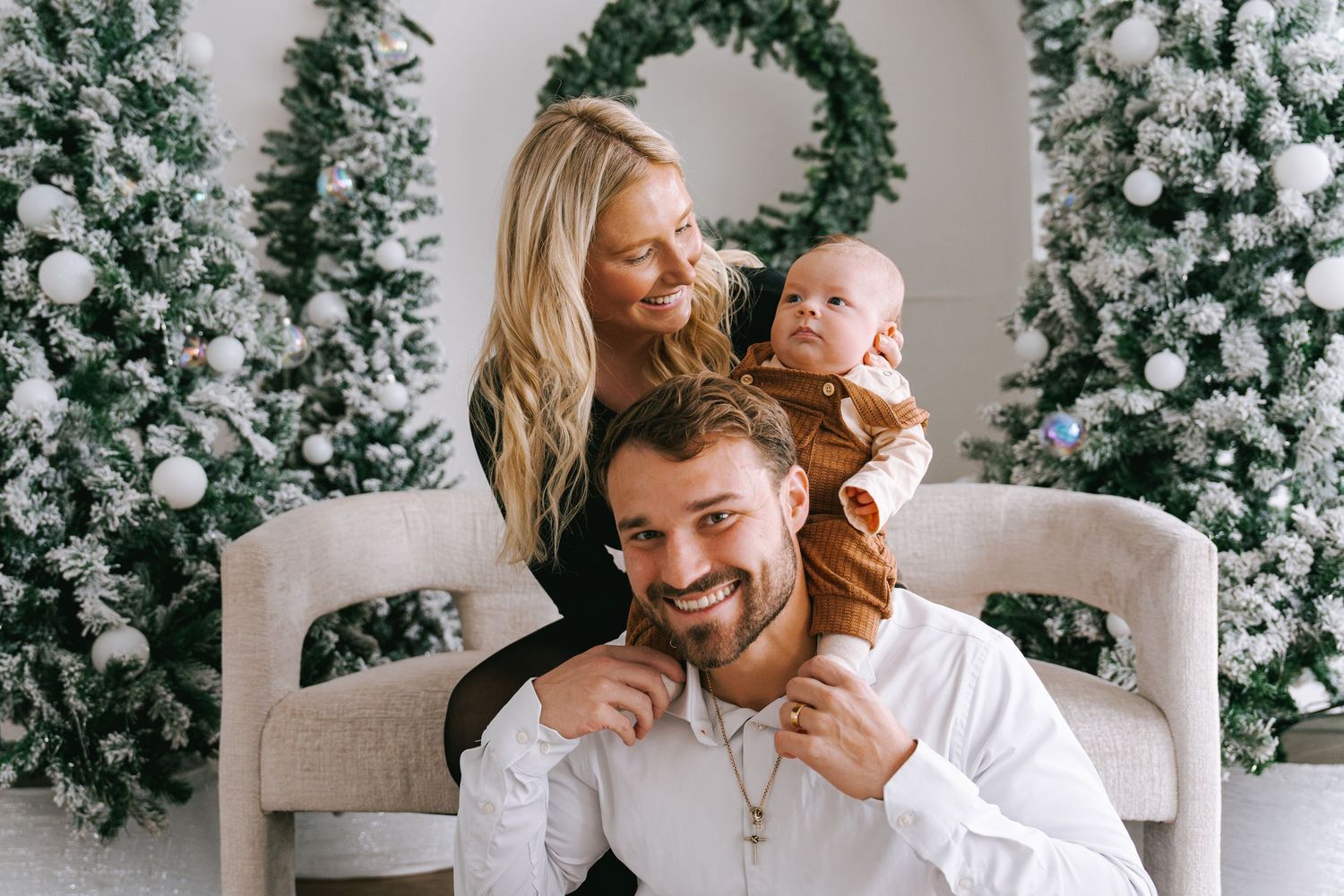 A family poses together for a holiday photo surrounded by snow-covered Christmas trees and a wreath.