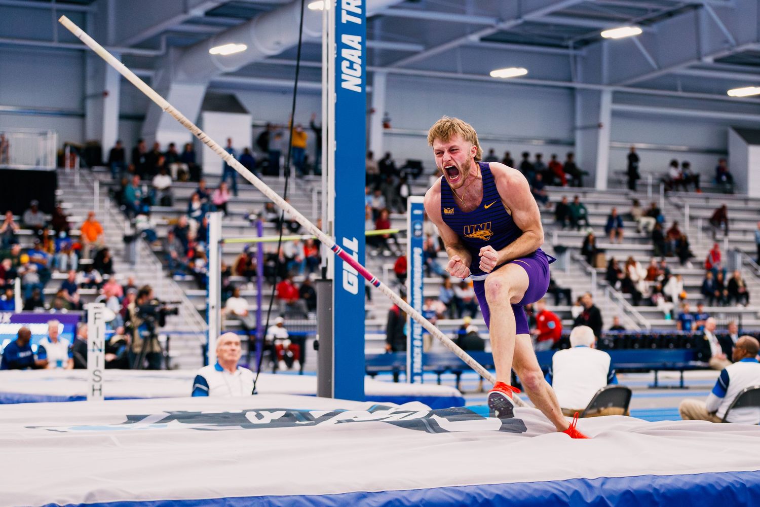 Athlete performs a pole vault attempt at an indoor track and field competition with spectators in the background.