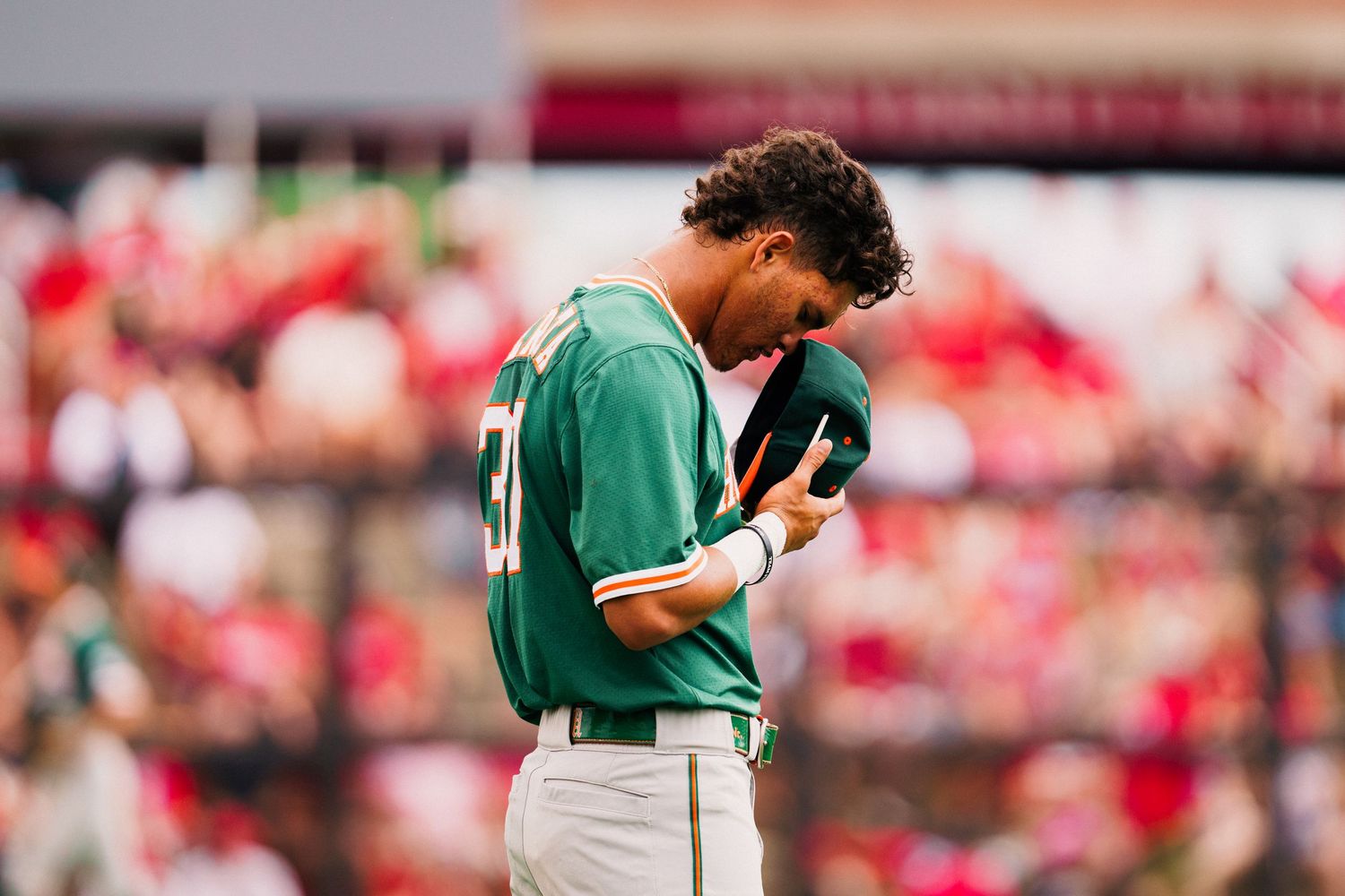 Baseball player in green jersey bows head during emotional moment at stadium with red background.