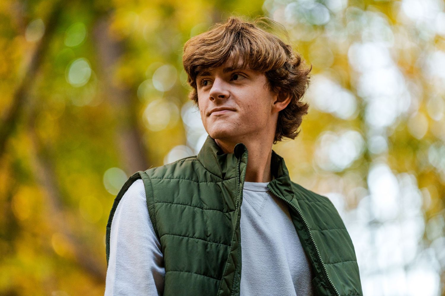 Person in green vest and white shirt poses against blurred fall foliage background.