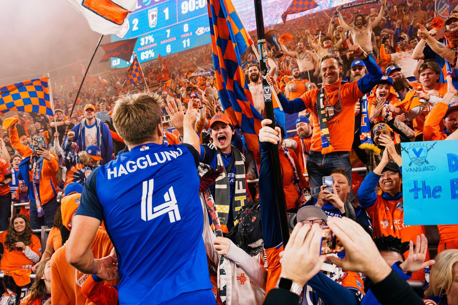 Basketball fans in orange and blue jerseys cheer and wave signs during an exciting game moment.
