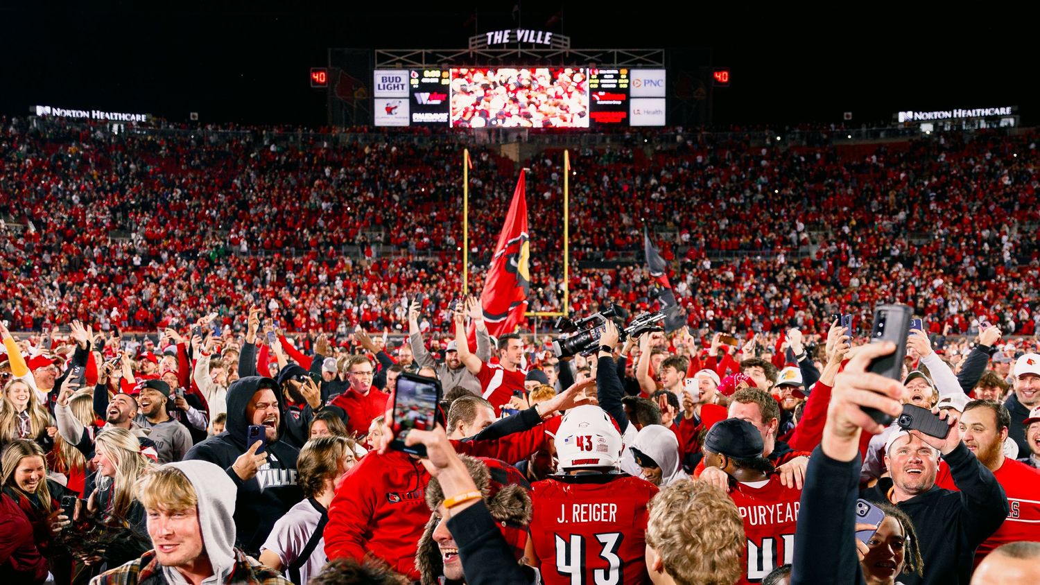 Excited crowd of Nebraska Cornhuskers fans celebrate during a night game at Memorial Stadium with red flags waving.
