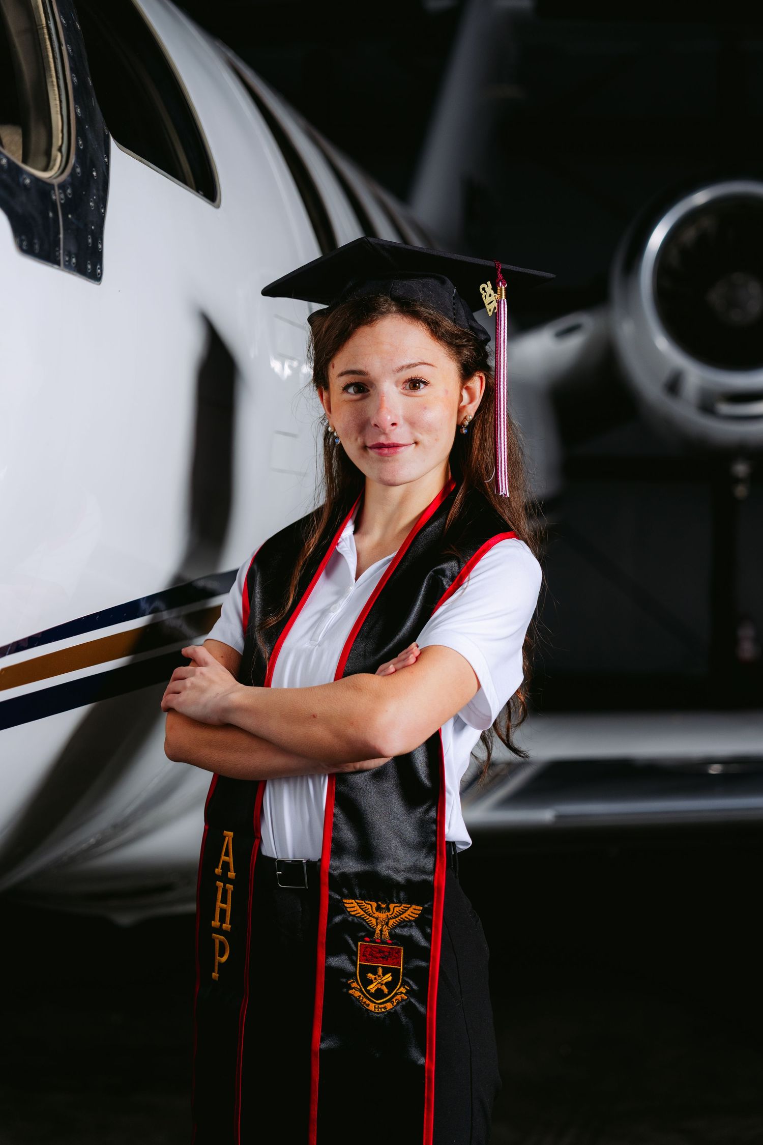 Graduate in cap and gown poses confidently in front of aircraft at aviation hangar for graduation photos.