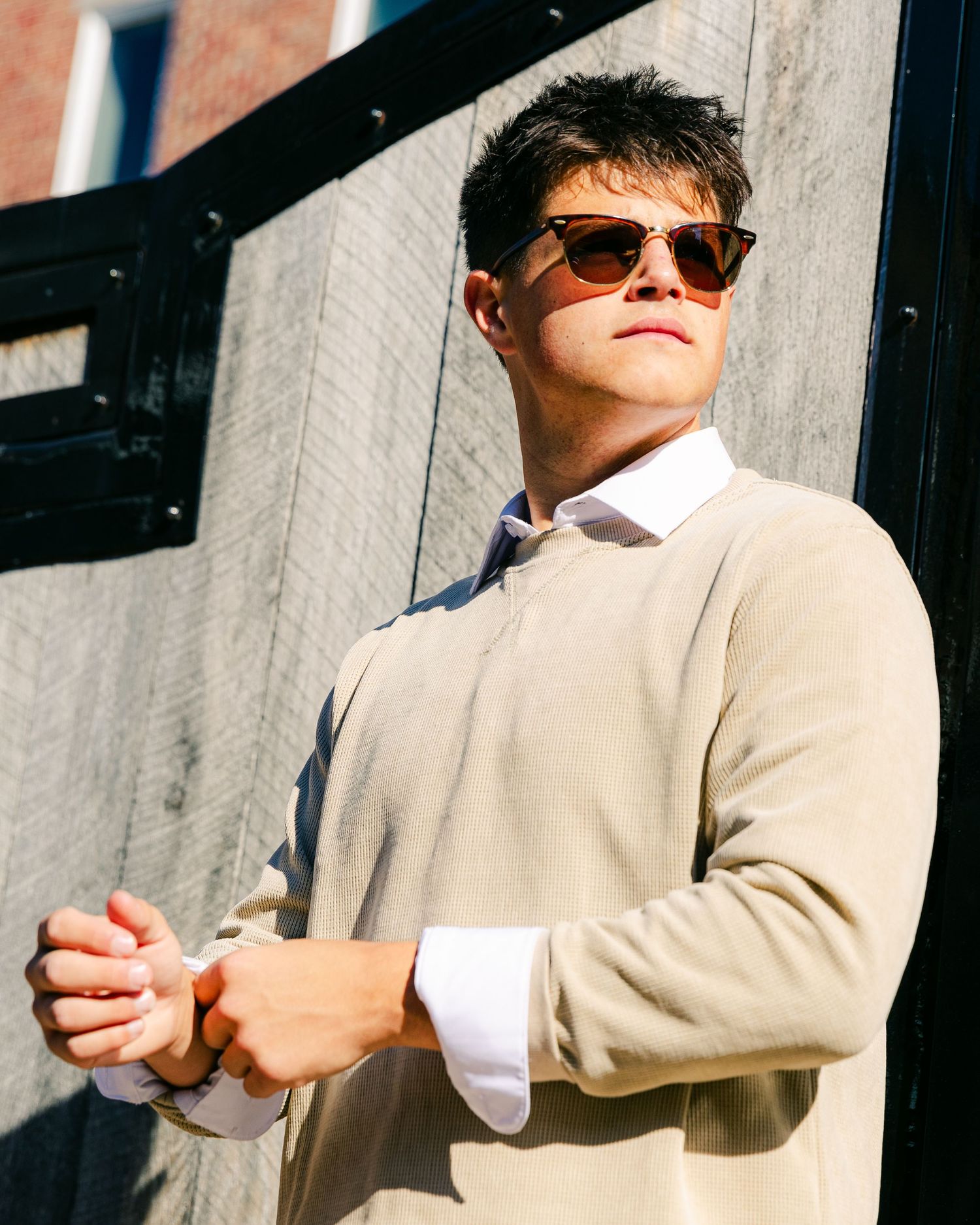 Young man in sunglasses and beige sweater leans against wall in dramatic sunlit portrait.