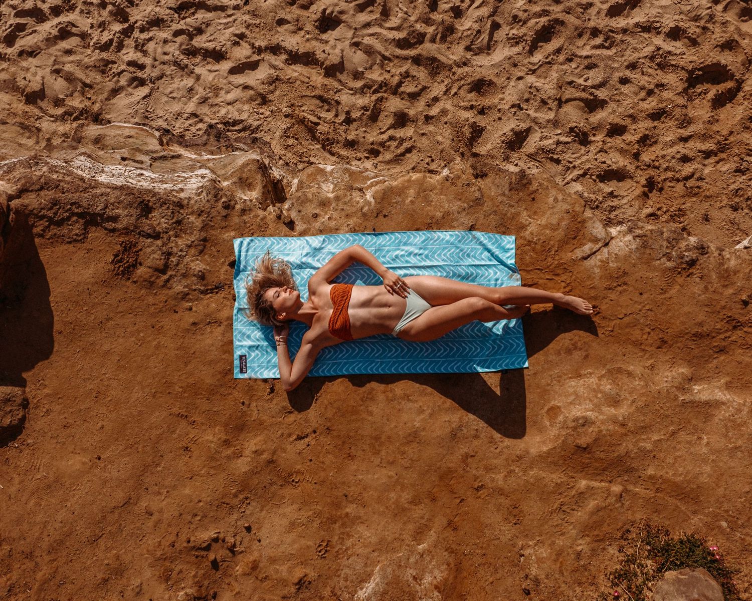 Aerial view of person lounging on blue towel against red cliff backdrop.