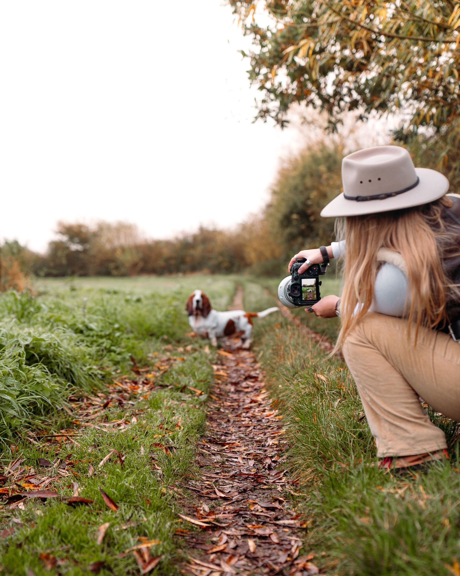 Photographer wearing tan vest and hat takes photos while lying on autumn ground covered in fallen leaves.