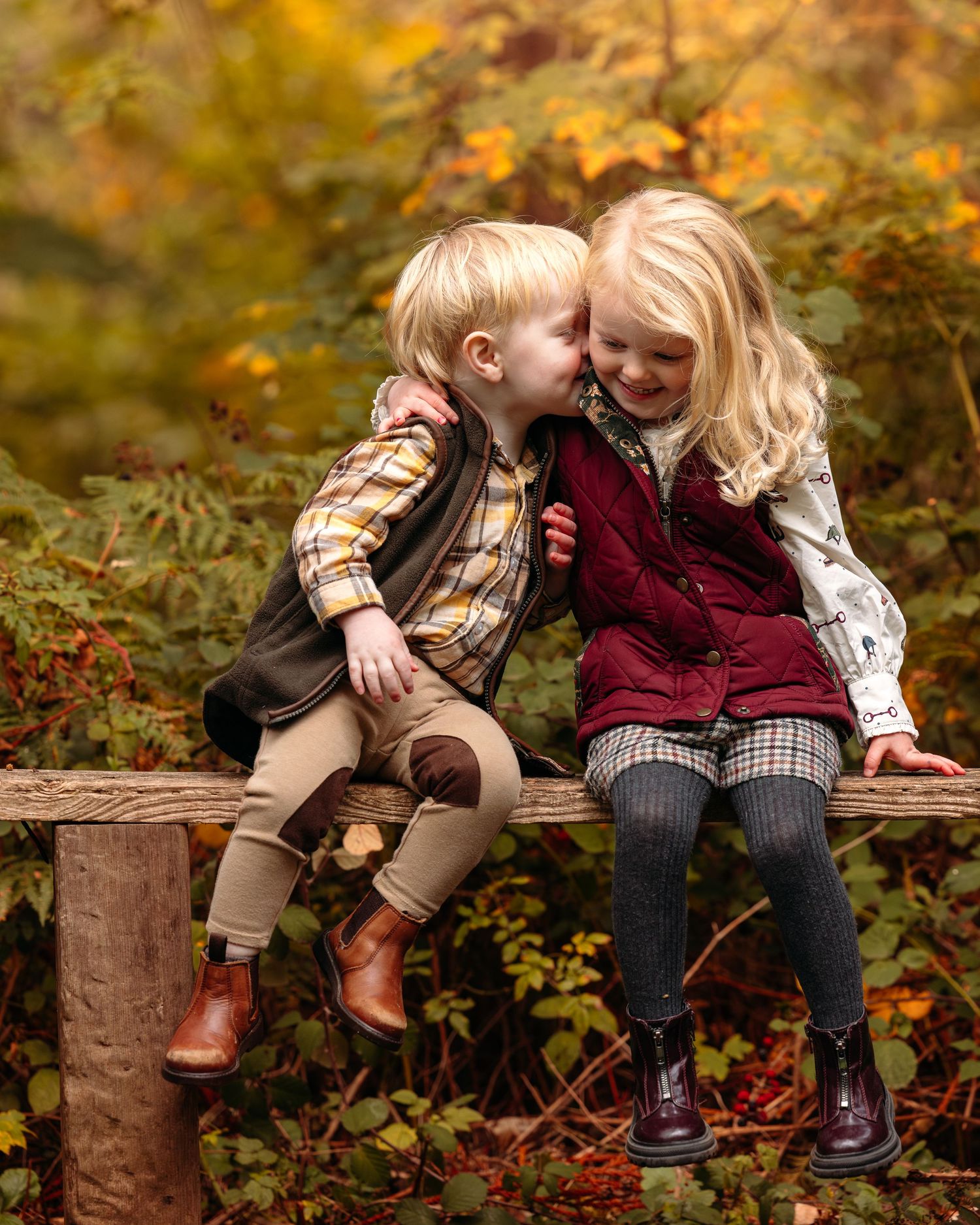 Children sitting on wooden logs surrounded by fall foliage wearing coordinated autumn outfits.