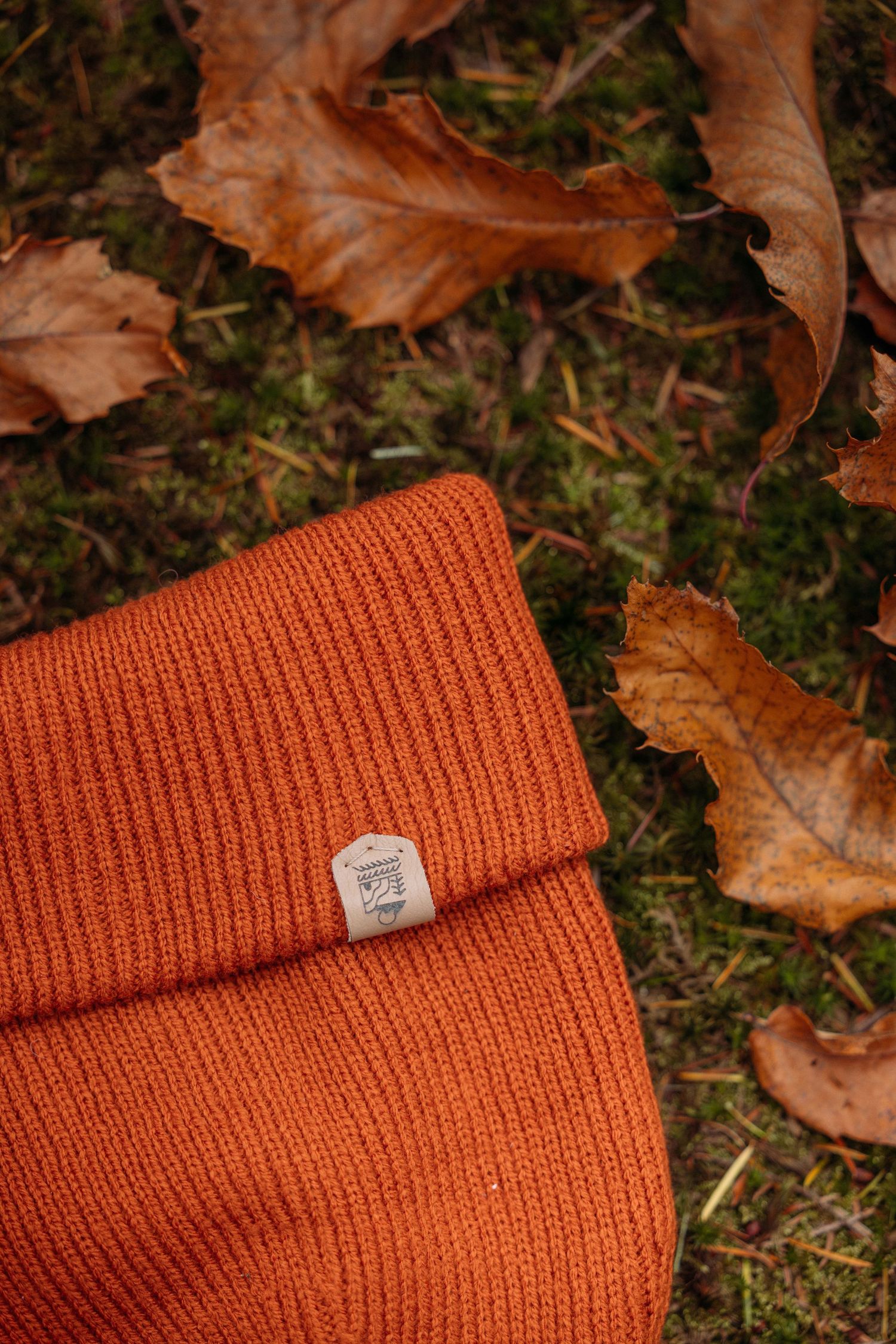 Orange knit beanie with brand tag lying on grass with autumn leaves.