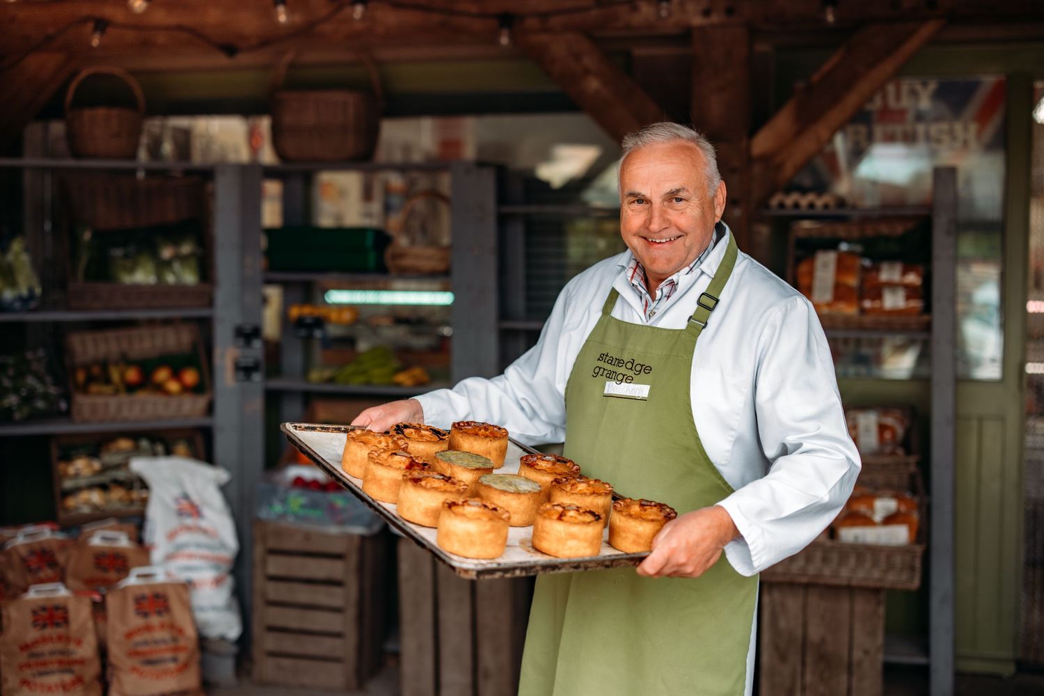 Baker in green apron displaying fresh baked goods in rustic store setting.