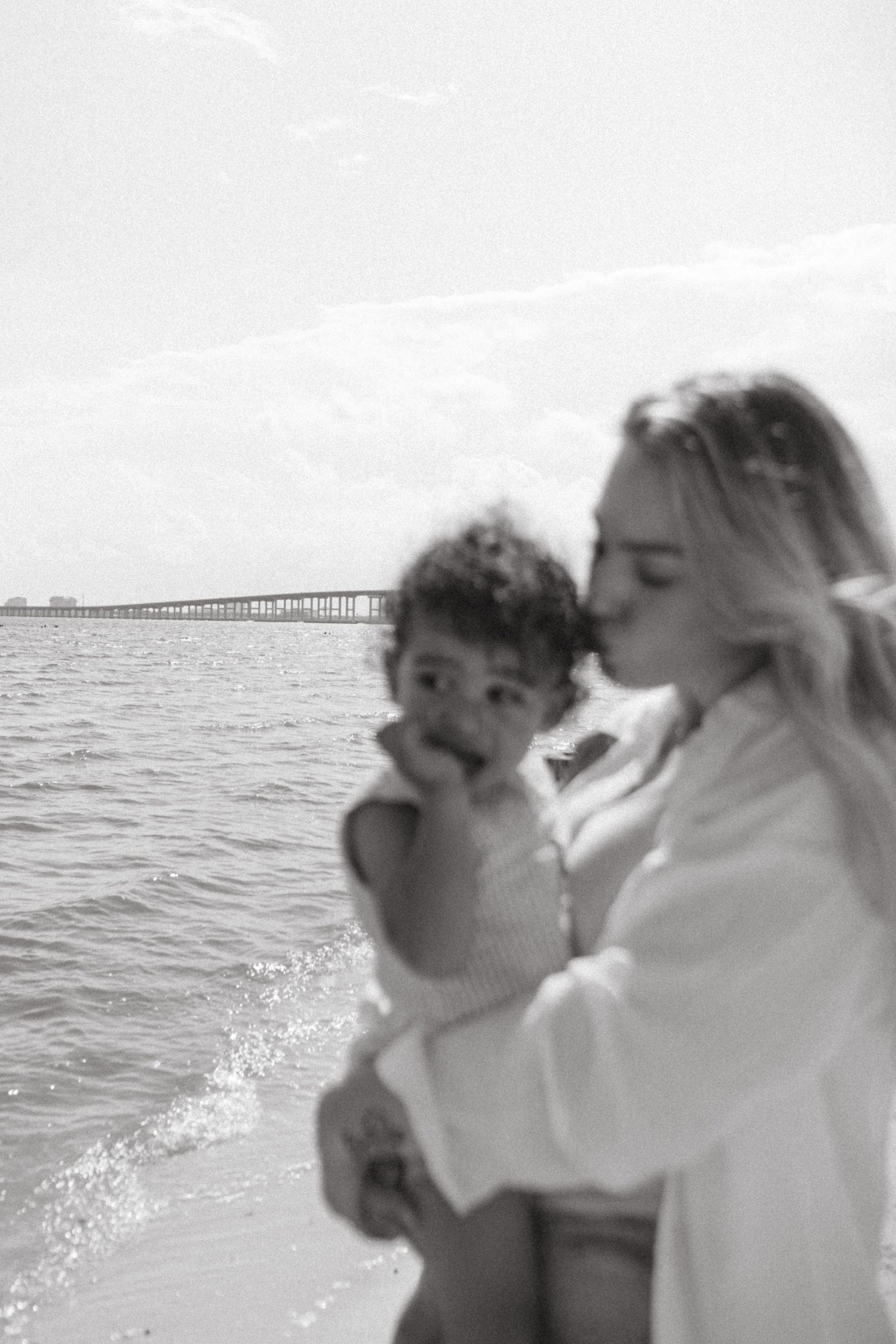 A tender black and white beach moment between parent and child near a pier on a bright sunny day.