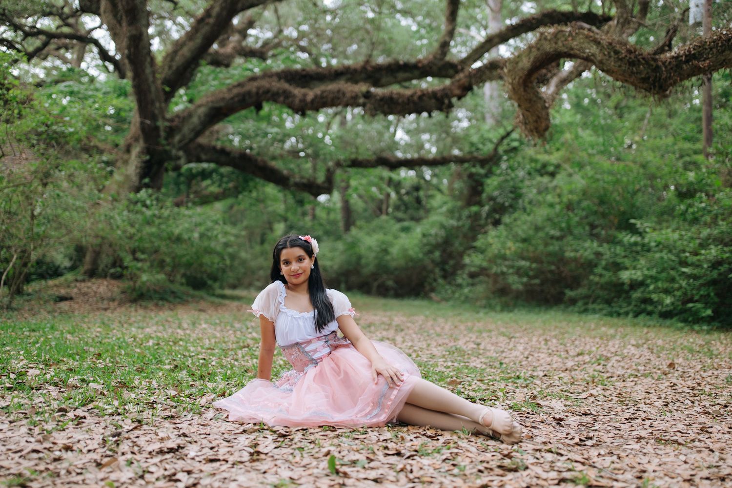 A ballet dancer in a pink tulle dress reclines gracefully beneath a sprawling oak tree in a wooded setting.