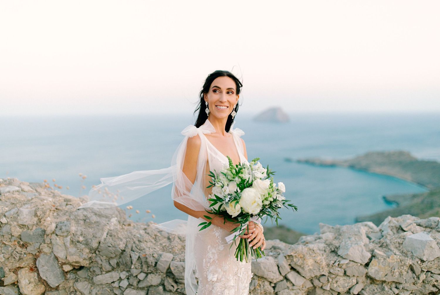 Elegant bride in lace dress holds white bouquet while standing on mountain cliff overlooking scenic water view.