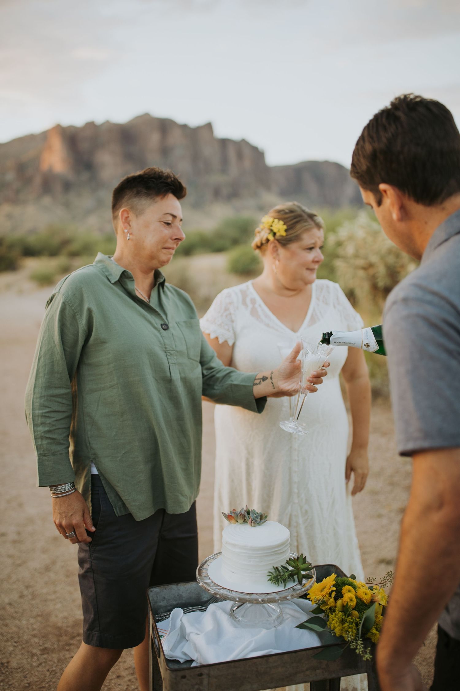 A desert wedding ceremony takes place with a small group gathered around a white cake against dramatic mountain backdrop.