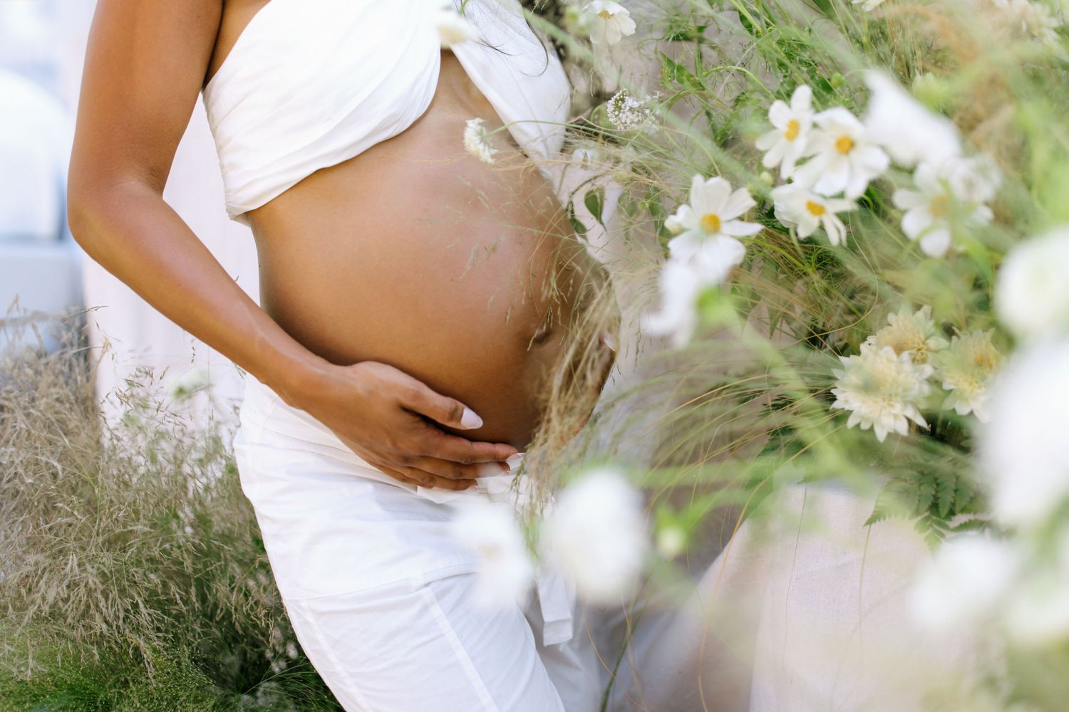 A maternity photoshoot captures delicate white flowers in focus with a blurred silhouette in white clothing in the background.