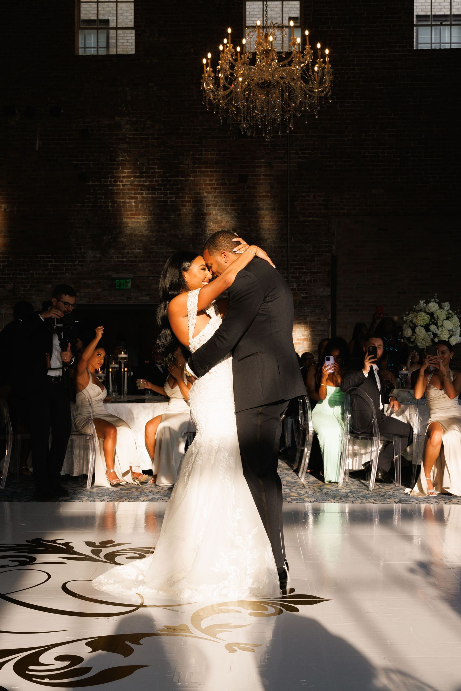 Newlyweds share their first dance under an elegant crystal chandelier on a black and white patterned dance floor.