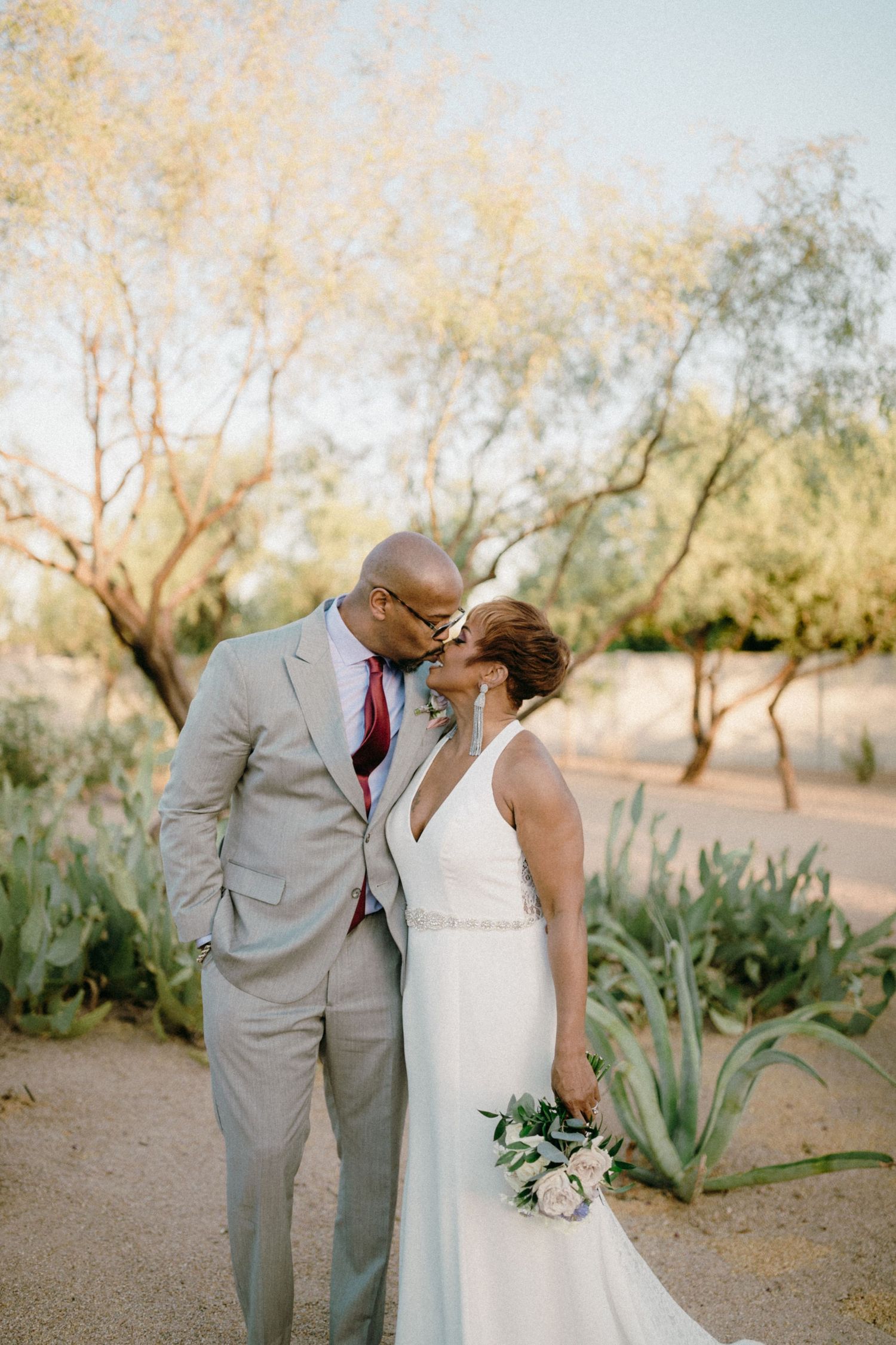 Newlyweds share an intimate moment outdoors in a desert garden setting with soft golden sunlight filtering through trees.