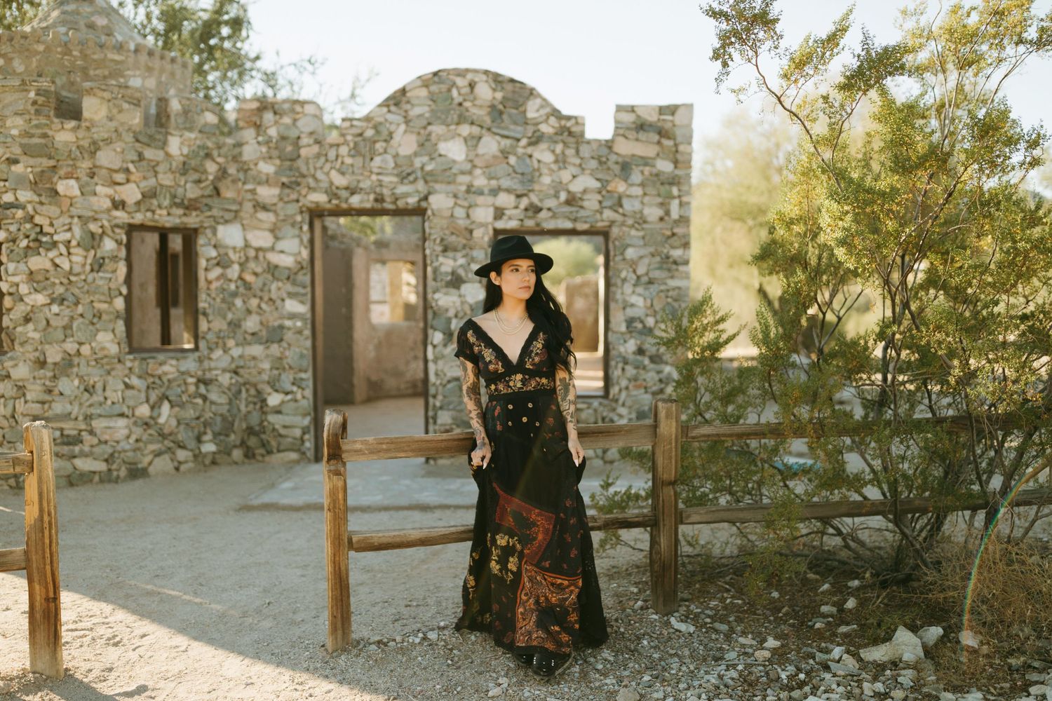 A person in a black dress and hat poses in front of a rustic stone building during golden hour.
