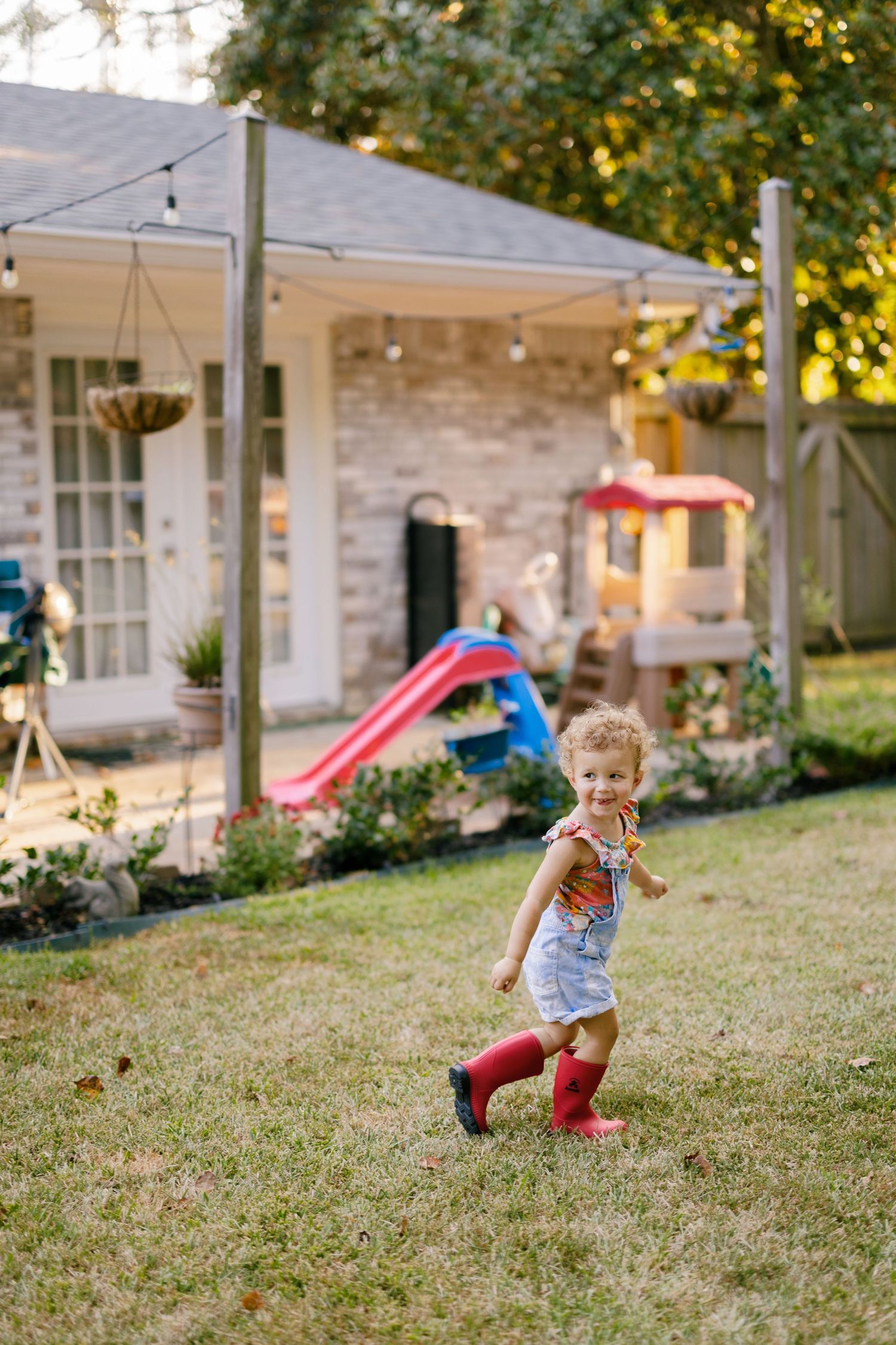 Kids run and play on a grassy slope in front of a brick house with string lights overhead.